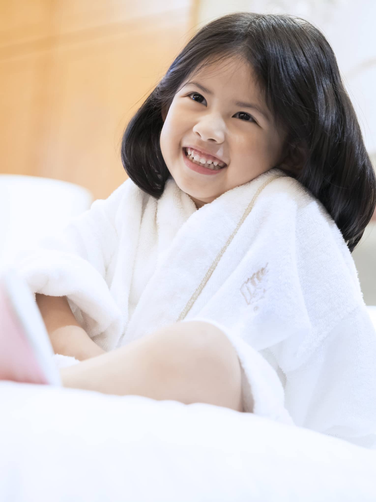 Smiling little girl wearing white bath robe and pink slippers sits on bed