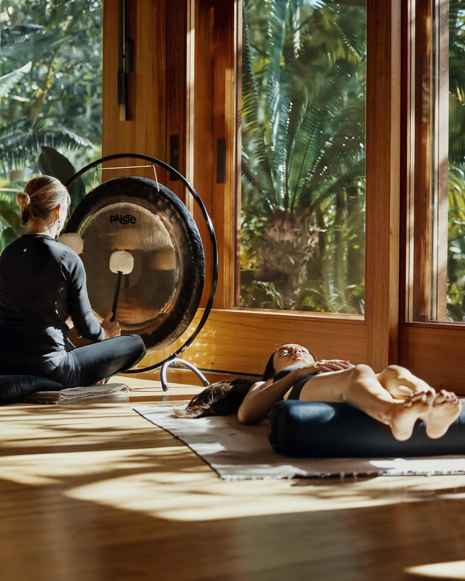 During a sound bath, a meditator lays on a mat, legs resting on a bolster, as a practitioner plays a gong with two mallets. 