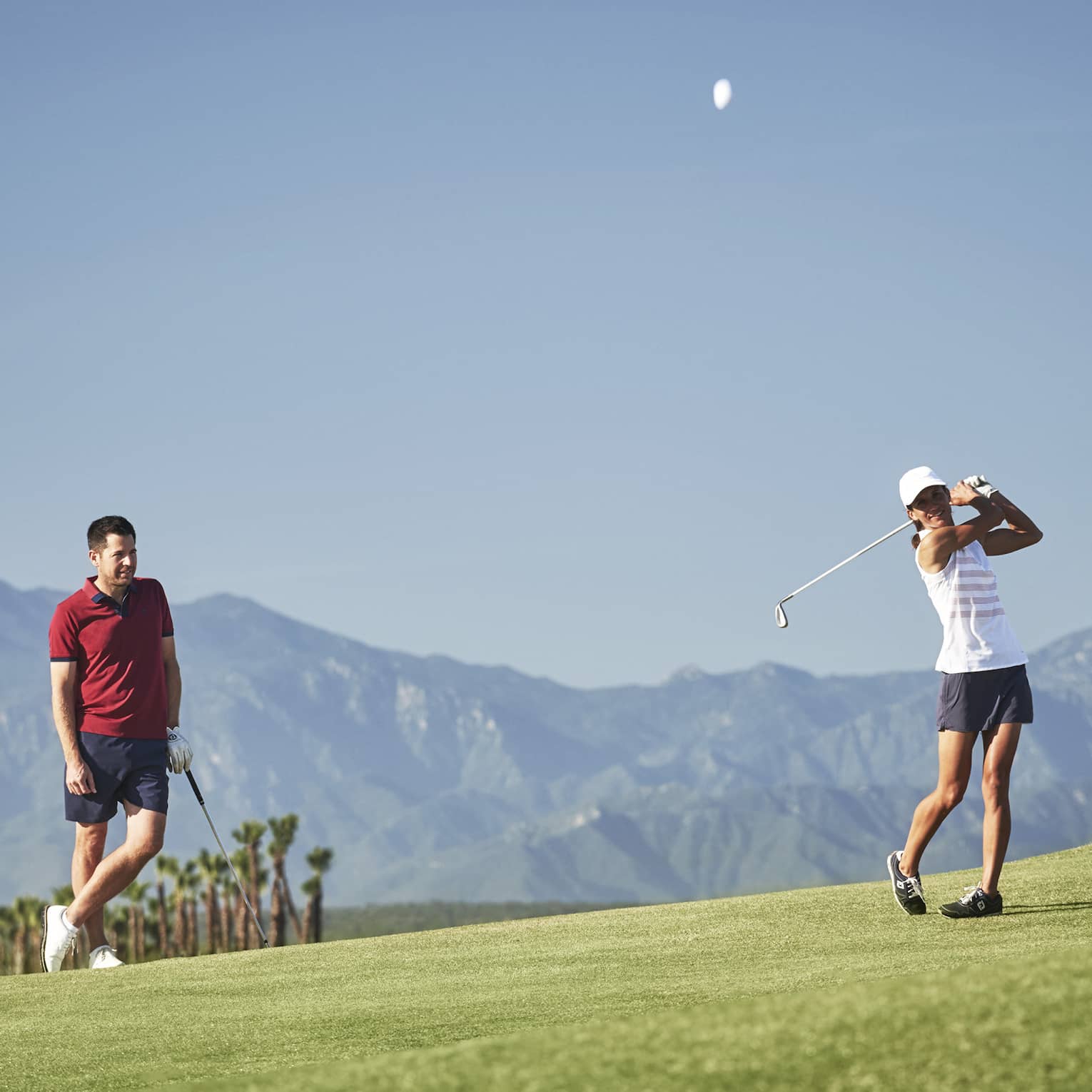 A golfer leans on their club observing another's swing on a picturesque golf course silhouetted by a mountain range.