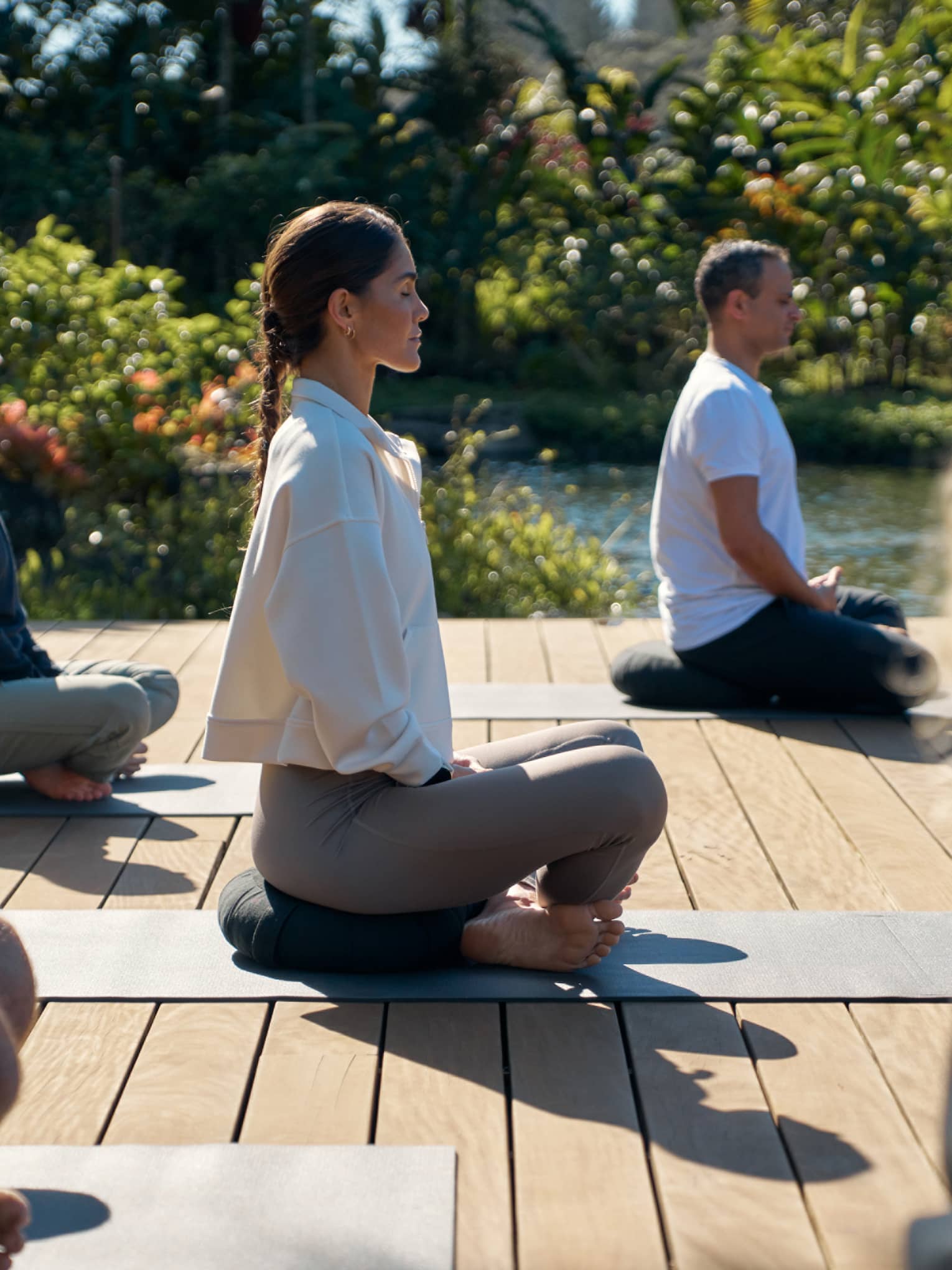 A group of people sit cross legged on wooden deck in garden setting, practicing yoga