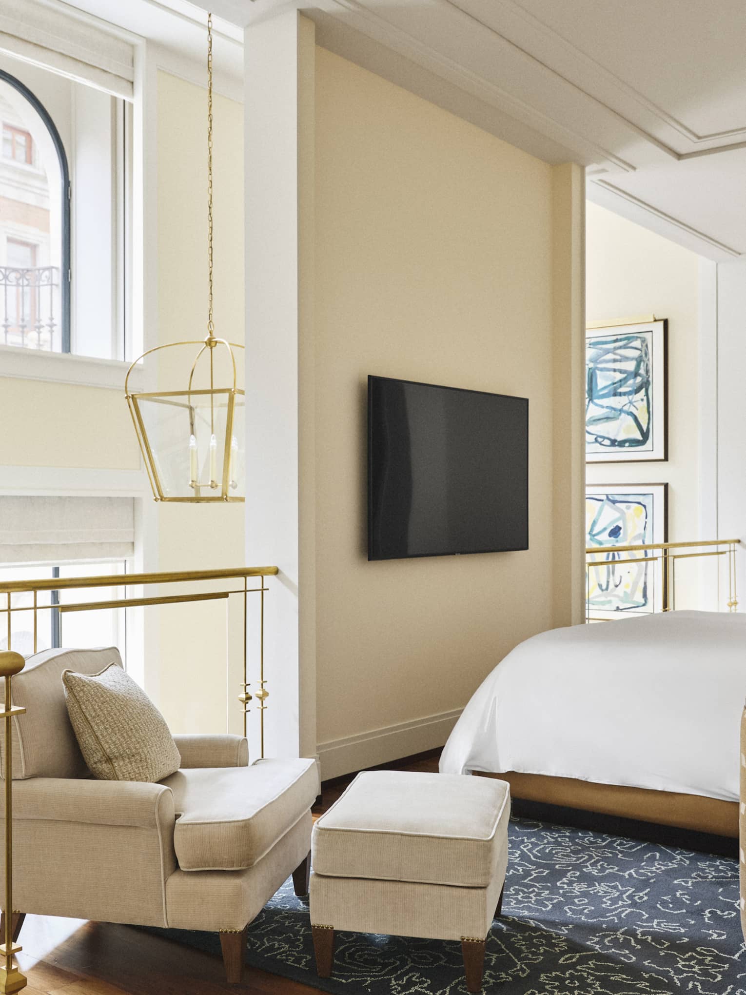Luxury hotel loft bedroom with a bed, wall-mounted TV, beige armchair and ottoman near a gold-railed balcony, hanging lantern-style lights and abstract wall art