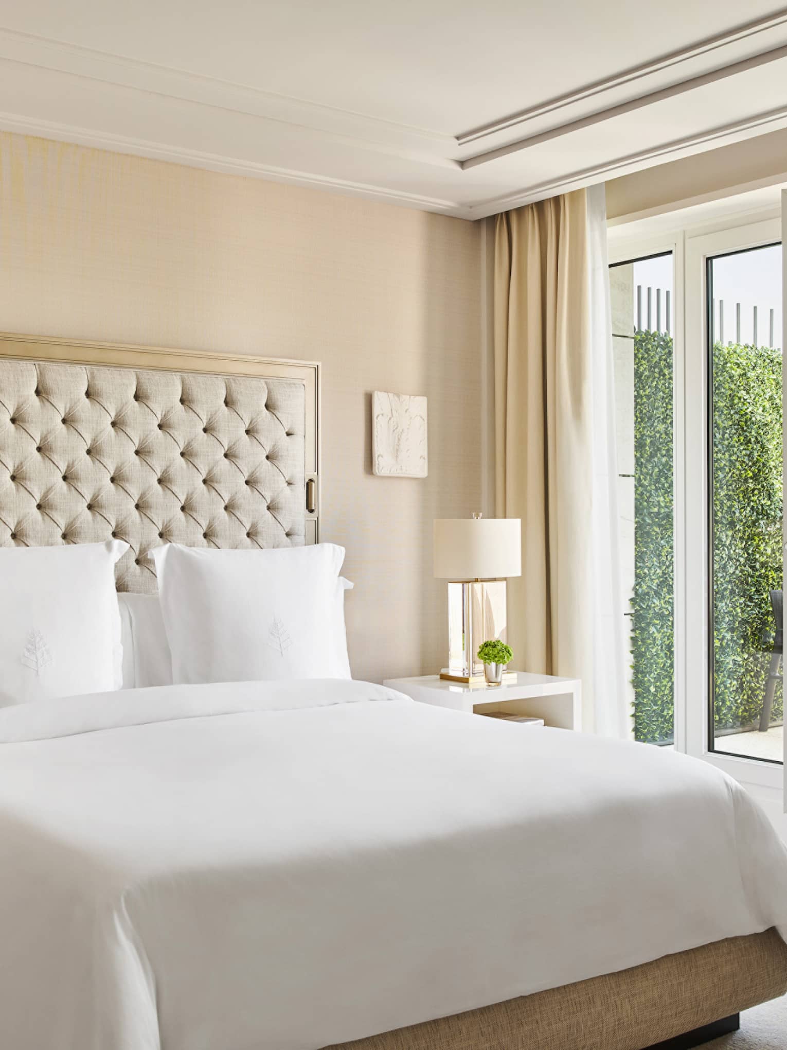 Luxury hotel bedroom with a king bed, tufted headboard, bedside tables with lamps and French doors opening to a private terrace with outdoor seating