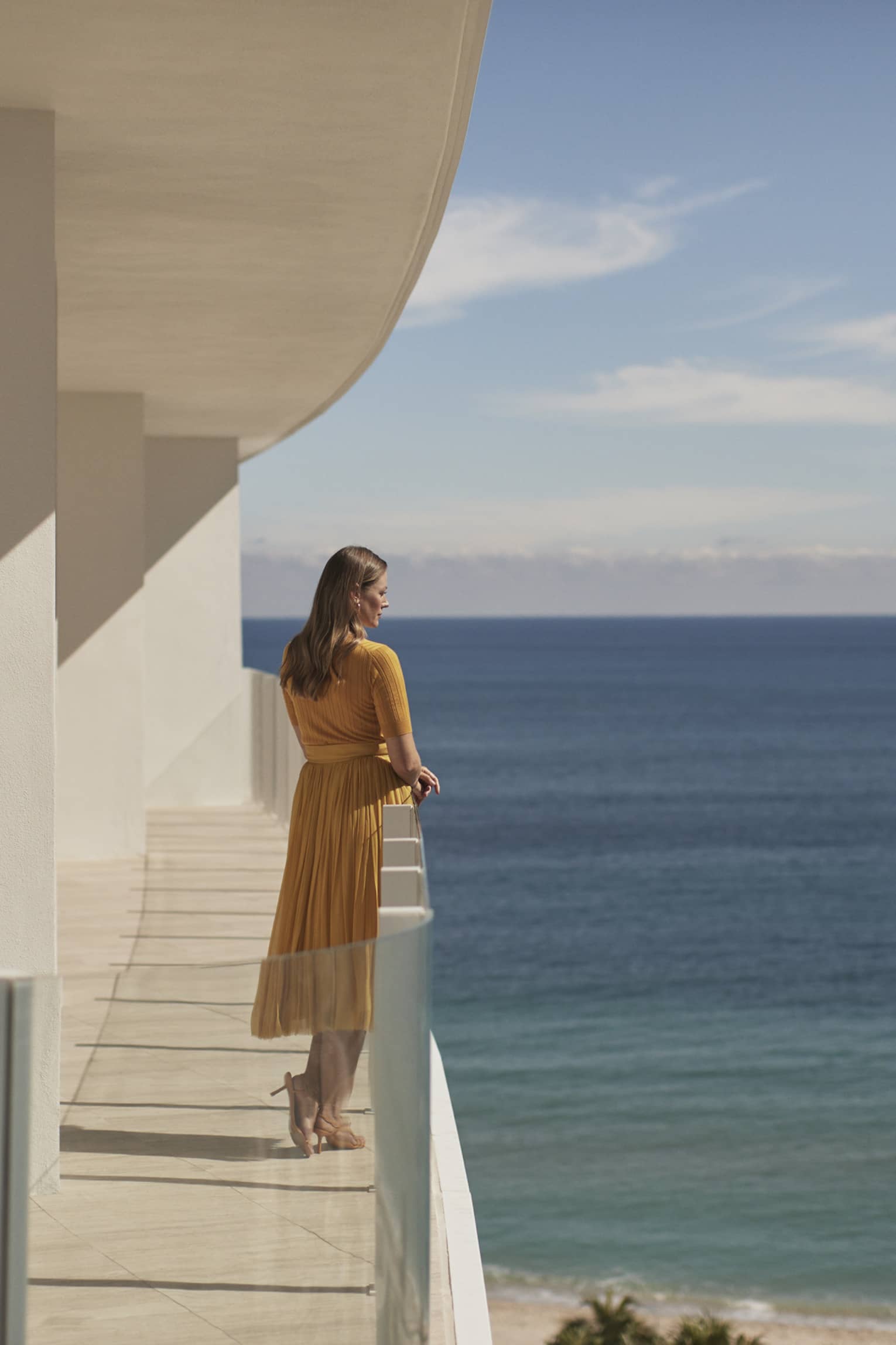 A woman standing on a balcony of a white stone building with the ocean in the background.
