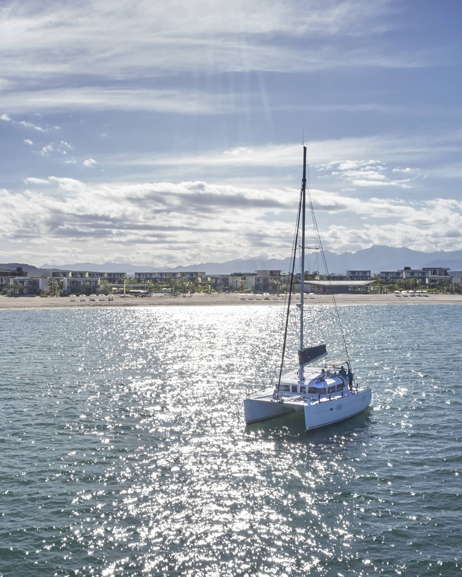 A trimaran boat sails over a glistening sea toward a shore lined with beach homes, distant mountains along the horizon.