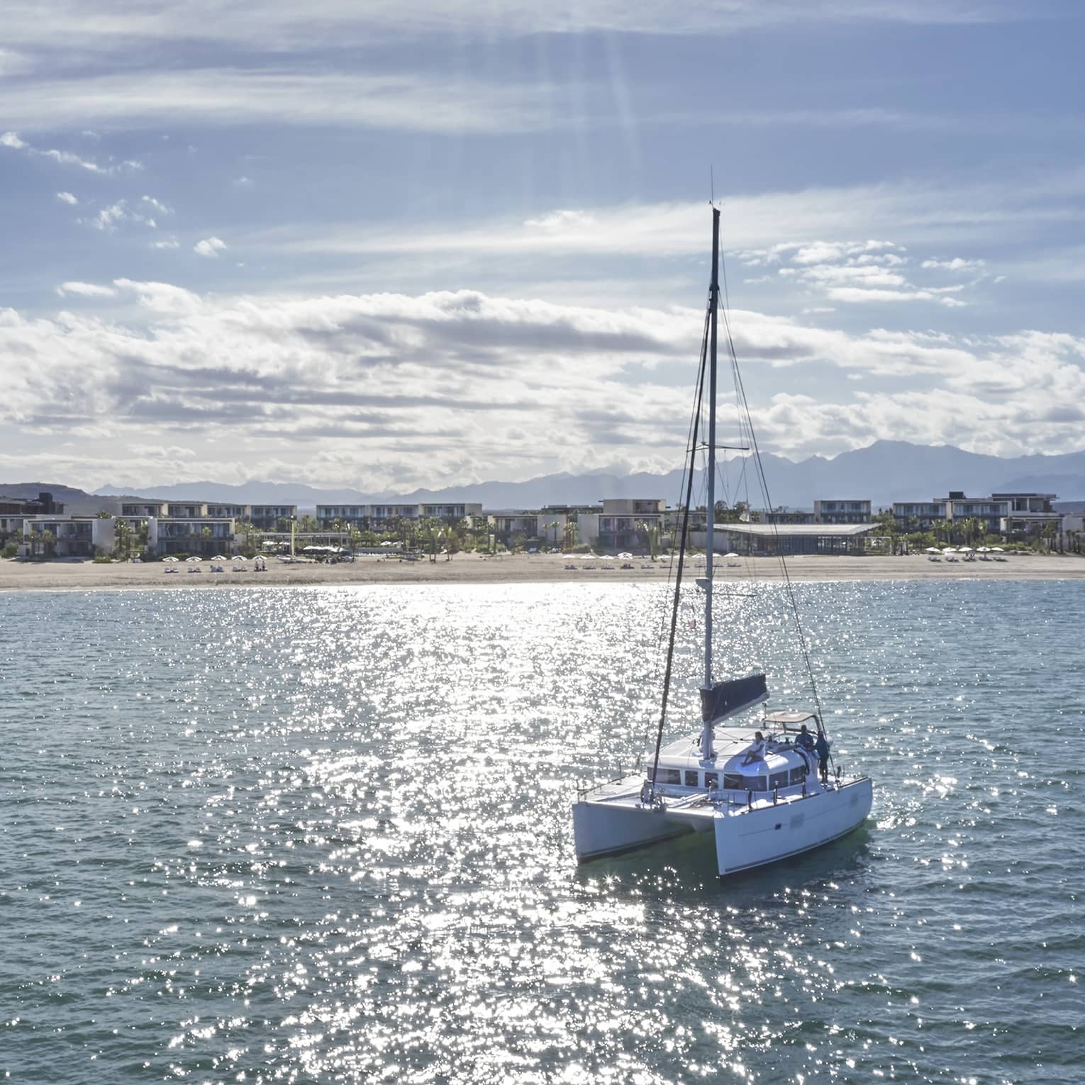 A trimaran boat sails over a glistening sea toward a shore lined with beach homes, distant mountains along the horizon.