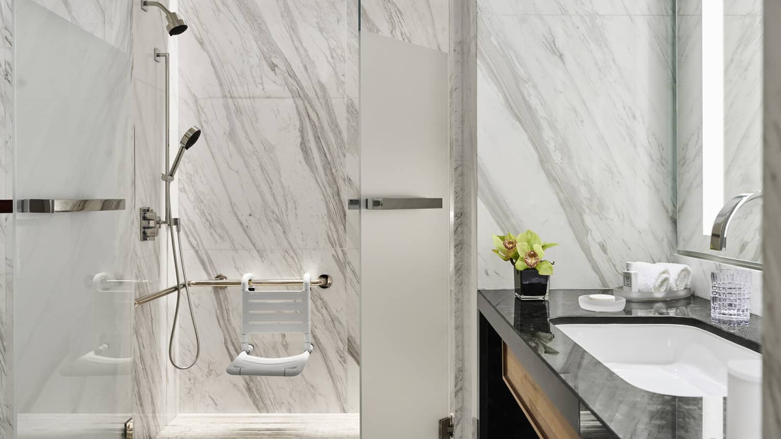 Luxurious marble bathroom with a glass-enclosed shower, shower seat, vanity with black countertop, chrome faucet, towels and a small flower arrangement