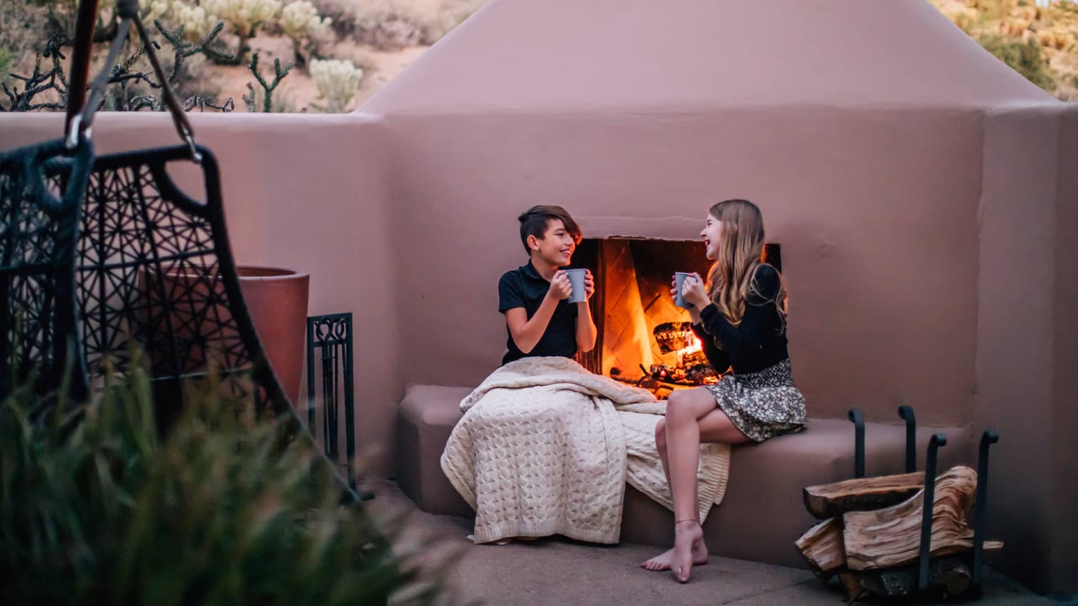 Two young guests sitting outside by the fire pit holding cups.