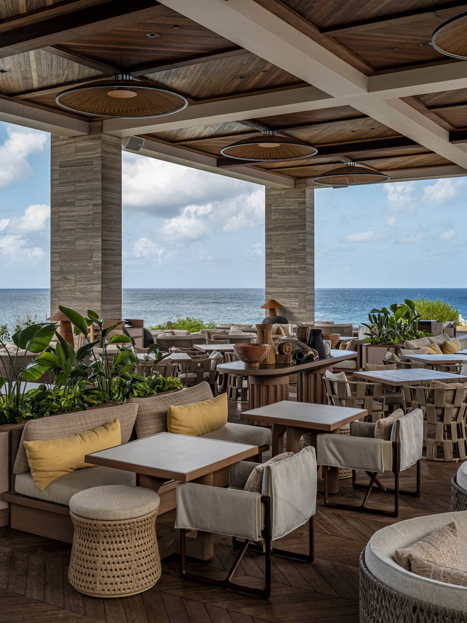 A spacious, covered outdoor dining space with small tables, comfortable chairs, plantings and ocean views