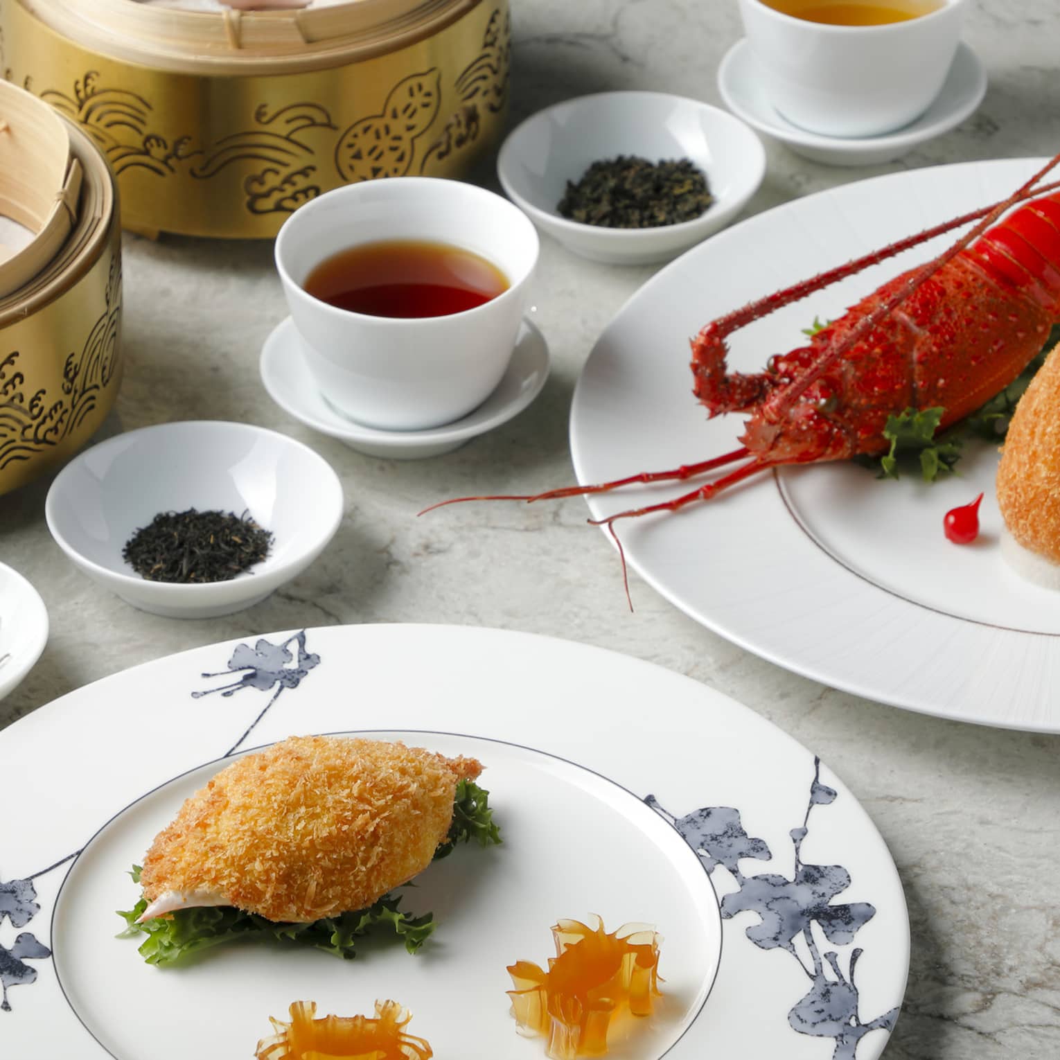 Plain white cups of tea are surrounded by bowls of dried tea leaves, baskets of dumplings and plates of lobster and seafood.