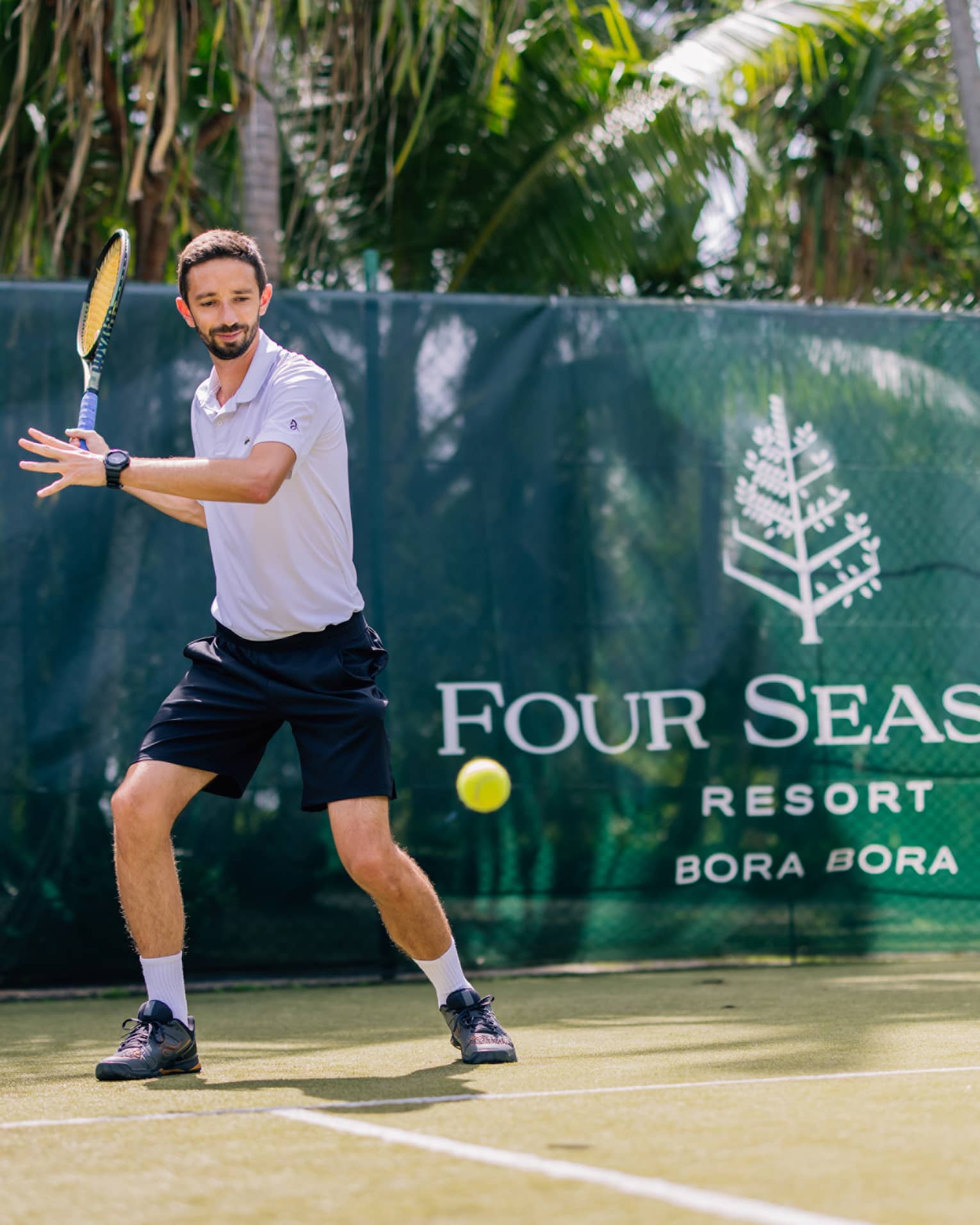 Standing behind the baseline on a court surrounded by palm trees, a tennis player prepares to hit a forehand.