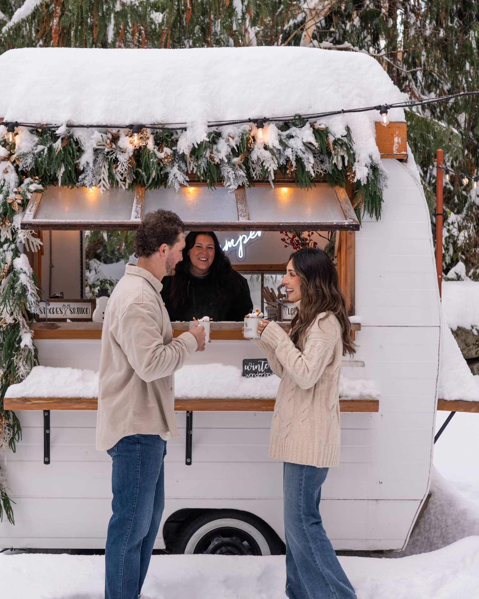 A couple holds mugs of hot chocolate outside of a camper in a snowy courtyard
