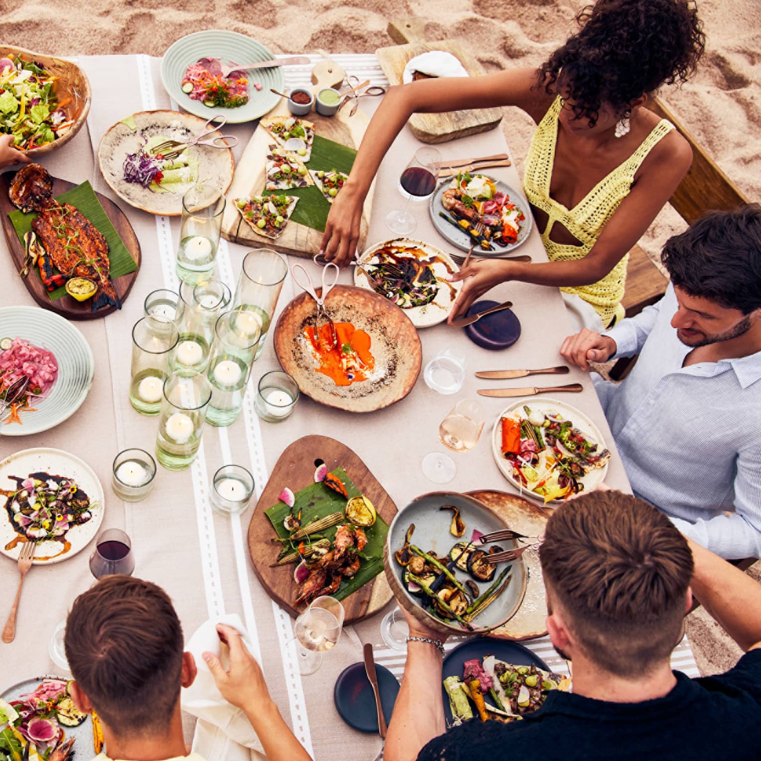 Beachside dining table with an assortment of vibrant dishes, candlelit glass holders and guests enjoying fresh seafood and grilled vegetables