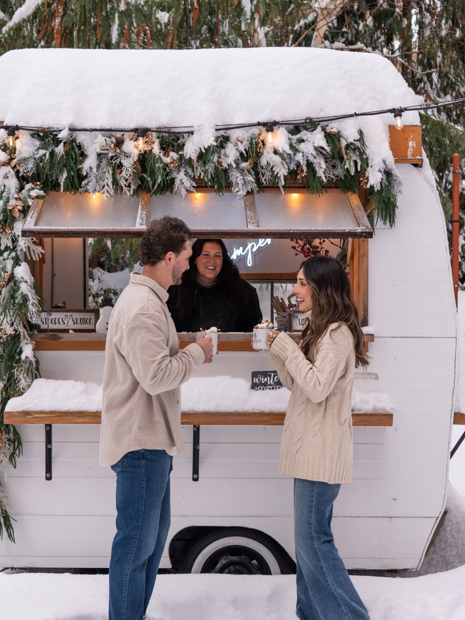 A couple holds mugs of hot chocolate outside of a camper in a snowy courtyard