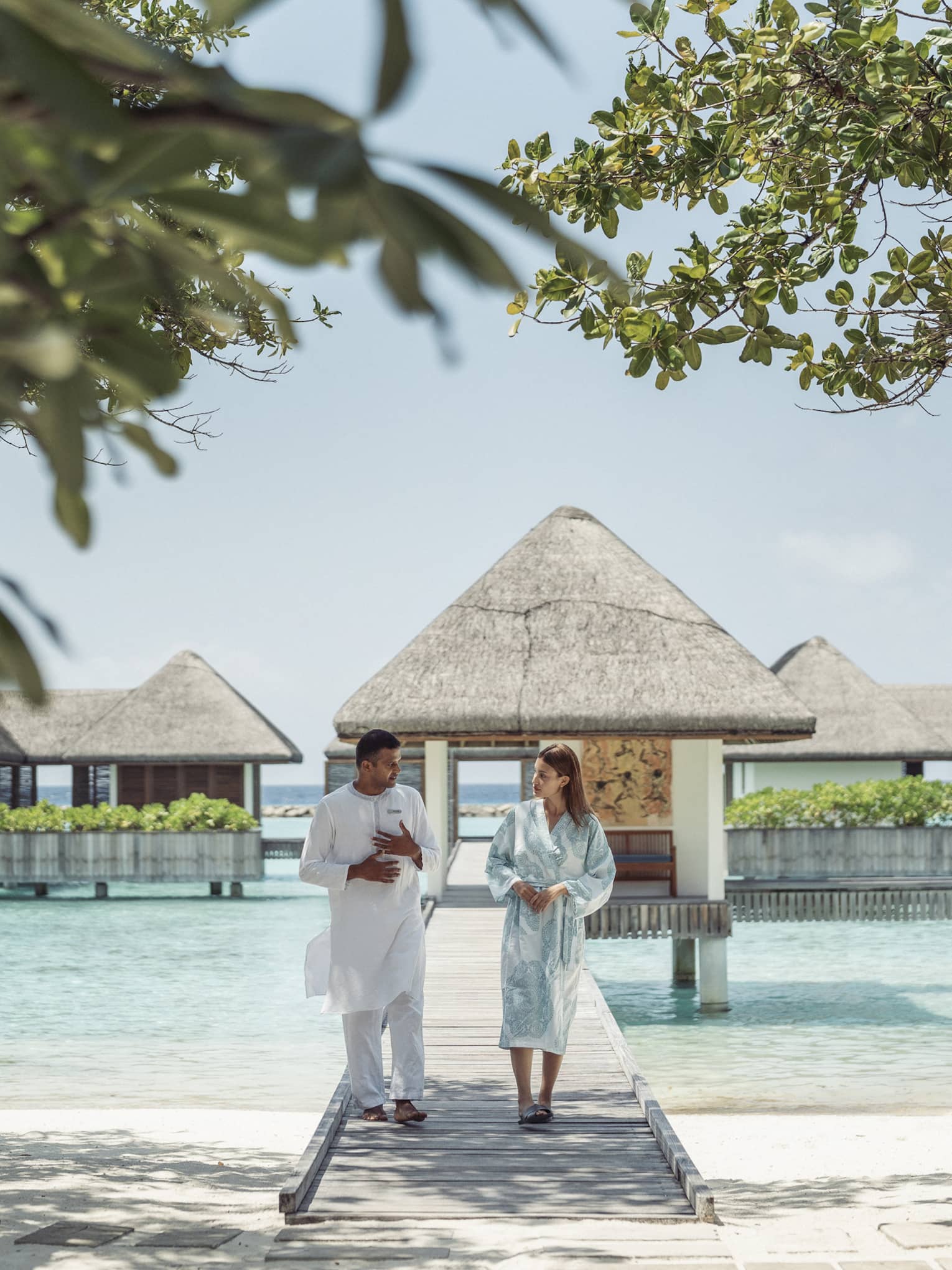 Two people walking along a wooden boardwalk in a wellness haven, surrounded by thatched-roof overwater villas and lush greenery in a tropical resort setting