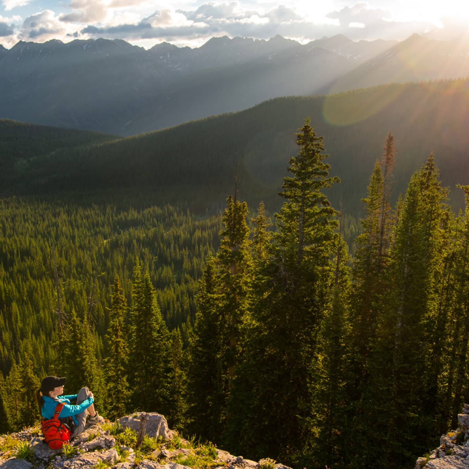 Person sits at edge of cliff overlooking trees, mountains as sun starts to set