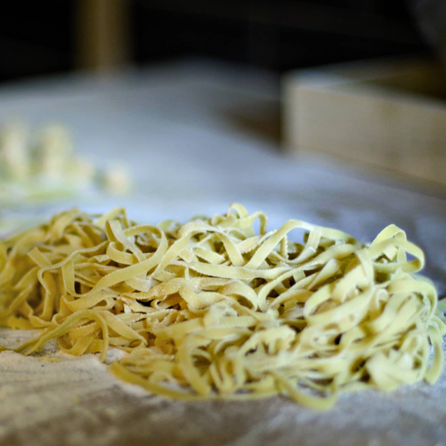 Pile of fresh tagliatelle rests on an aged wood counter covered with flour, beside scraps of pasta dough. 