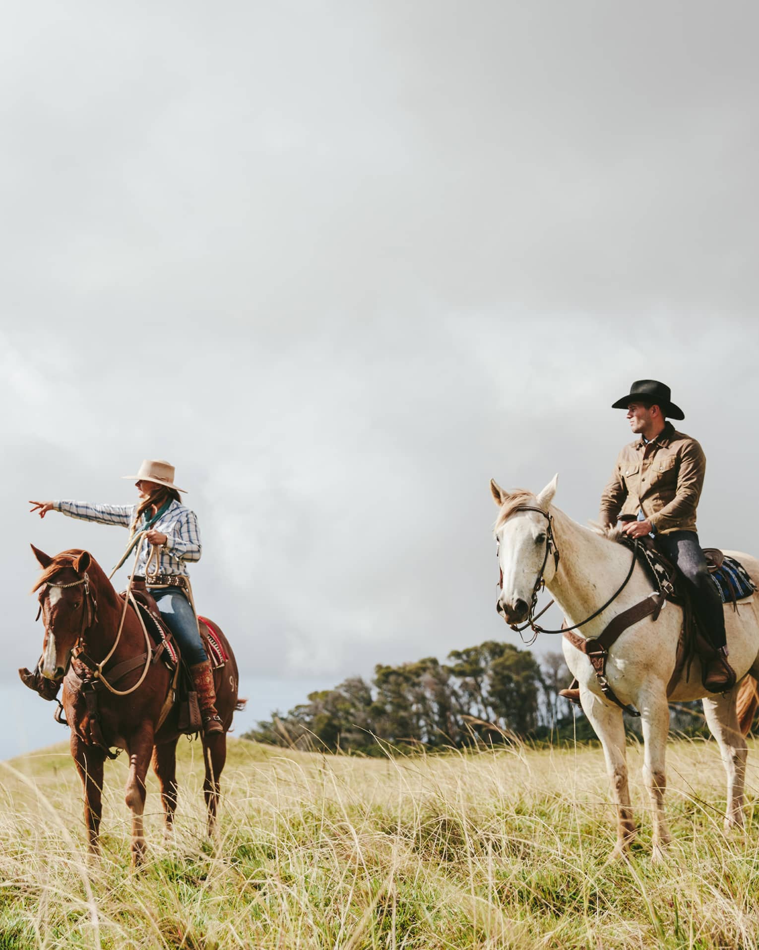 A couple riding horses, looking out at the long grass fields, with grassy hills and trees in the distance on a cloudy day.