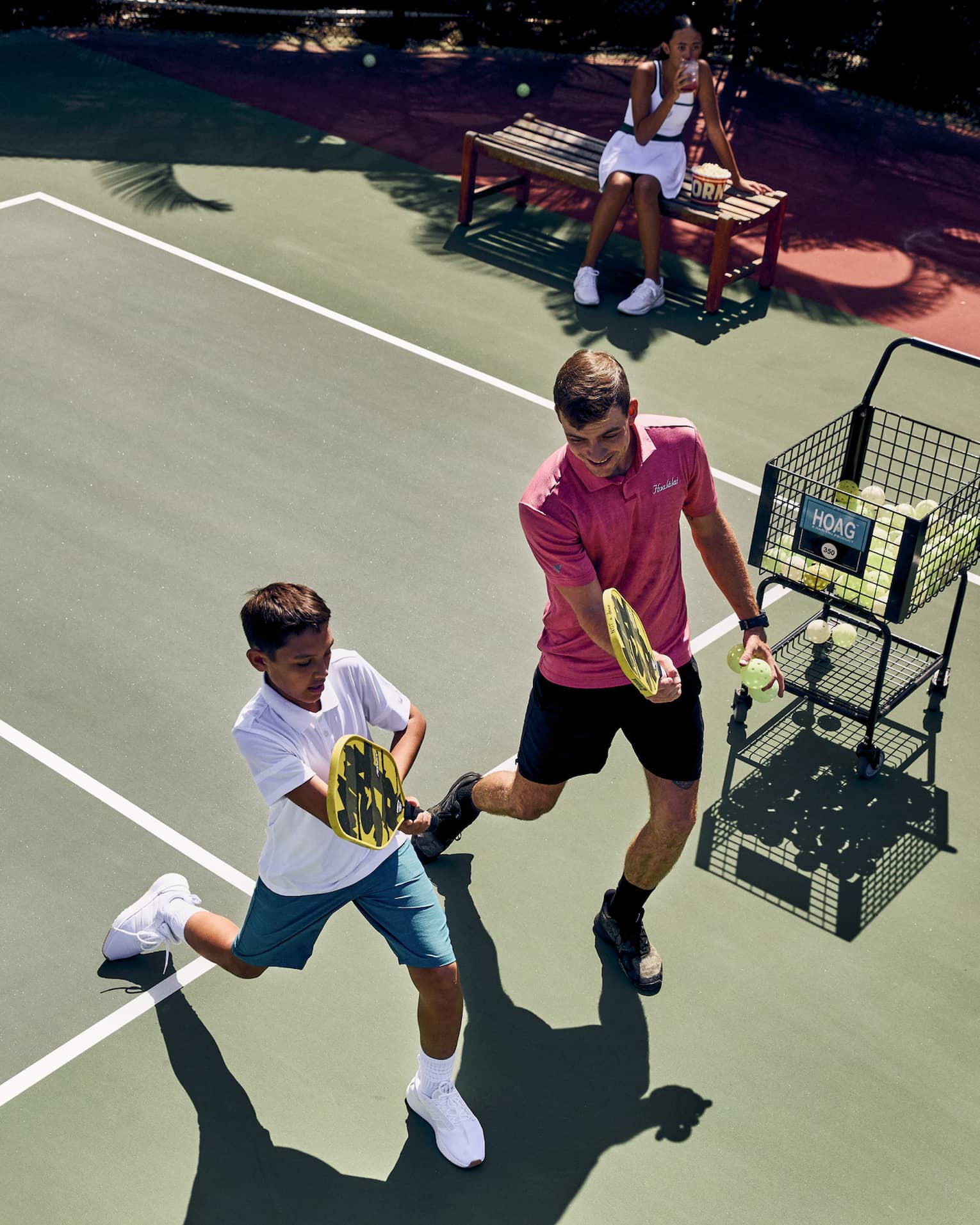 Aerial view of a pickleball pro instructing a player, both holding paddles while lunging forward, ball cart off to the side.