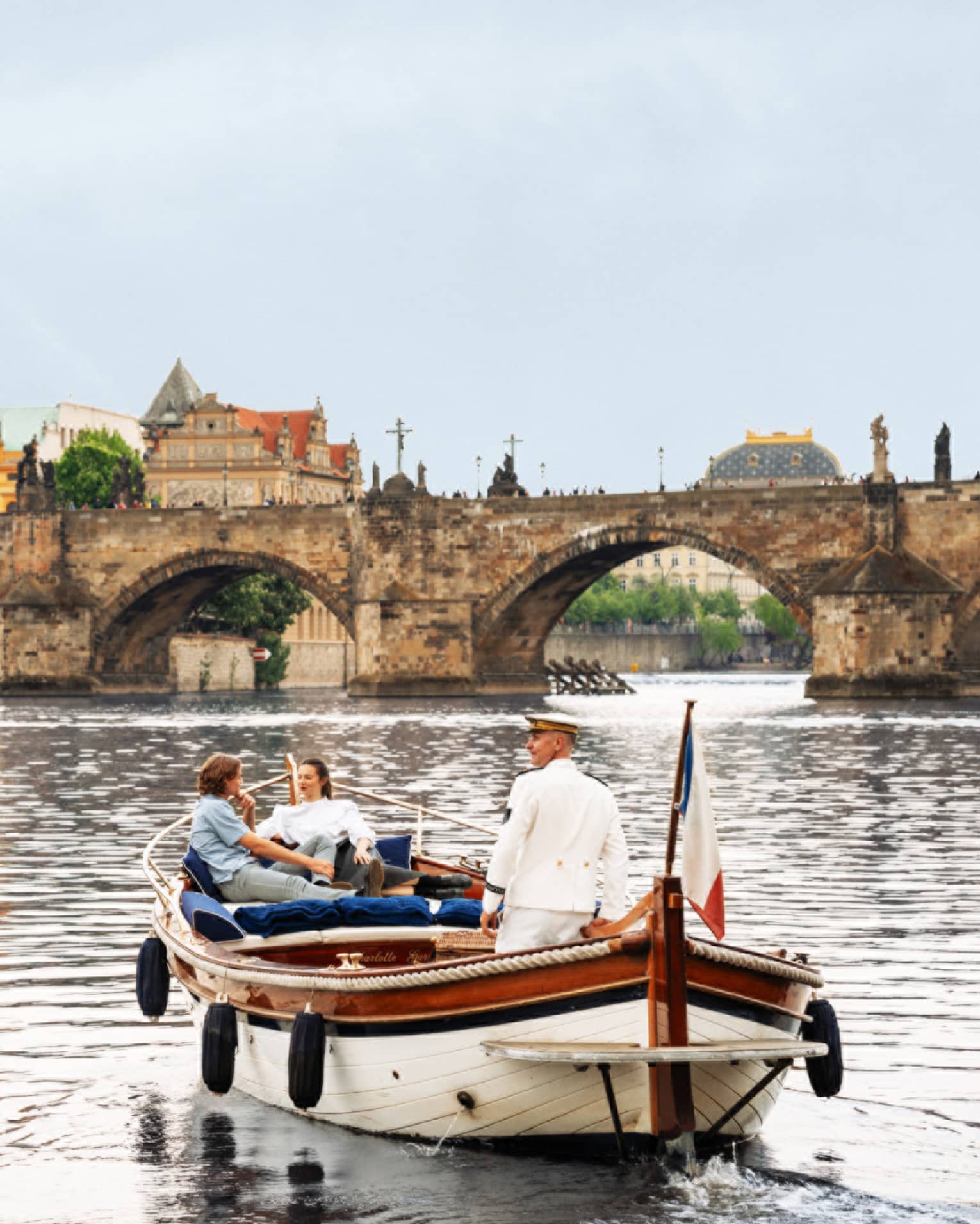Couple with captain in wooden boat on Vltava River with bridge and buildings in backdrop