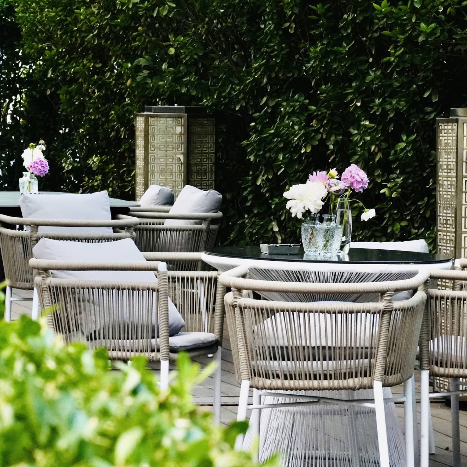Flanked by tall hedges, two tables surrounded by beige, cushioned rattan chairs hold glass vases of pink and white flowers.