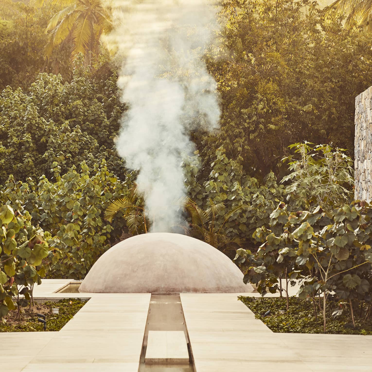 White steam rises from a small cement dome nestled between two large stone walls and surrounded by dense dark-green foliage.