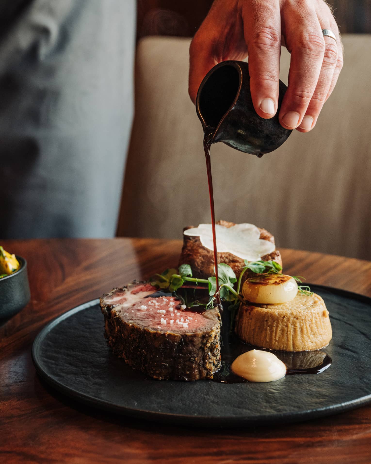 A waiter's hands pour sauce onto a slice of braised short rib served on a black plate next to two small black side bowls