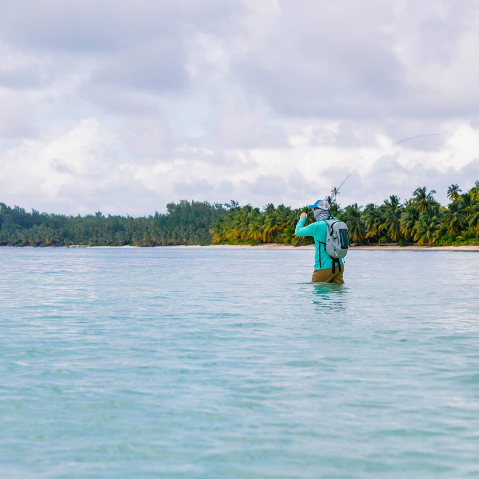 Long view of fly fisher in thigh-high water, face covered and rod held overhead in a backcast, forested beach island beyond. 