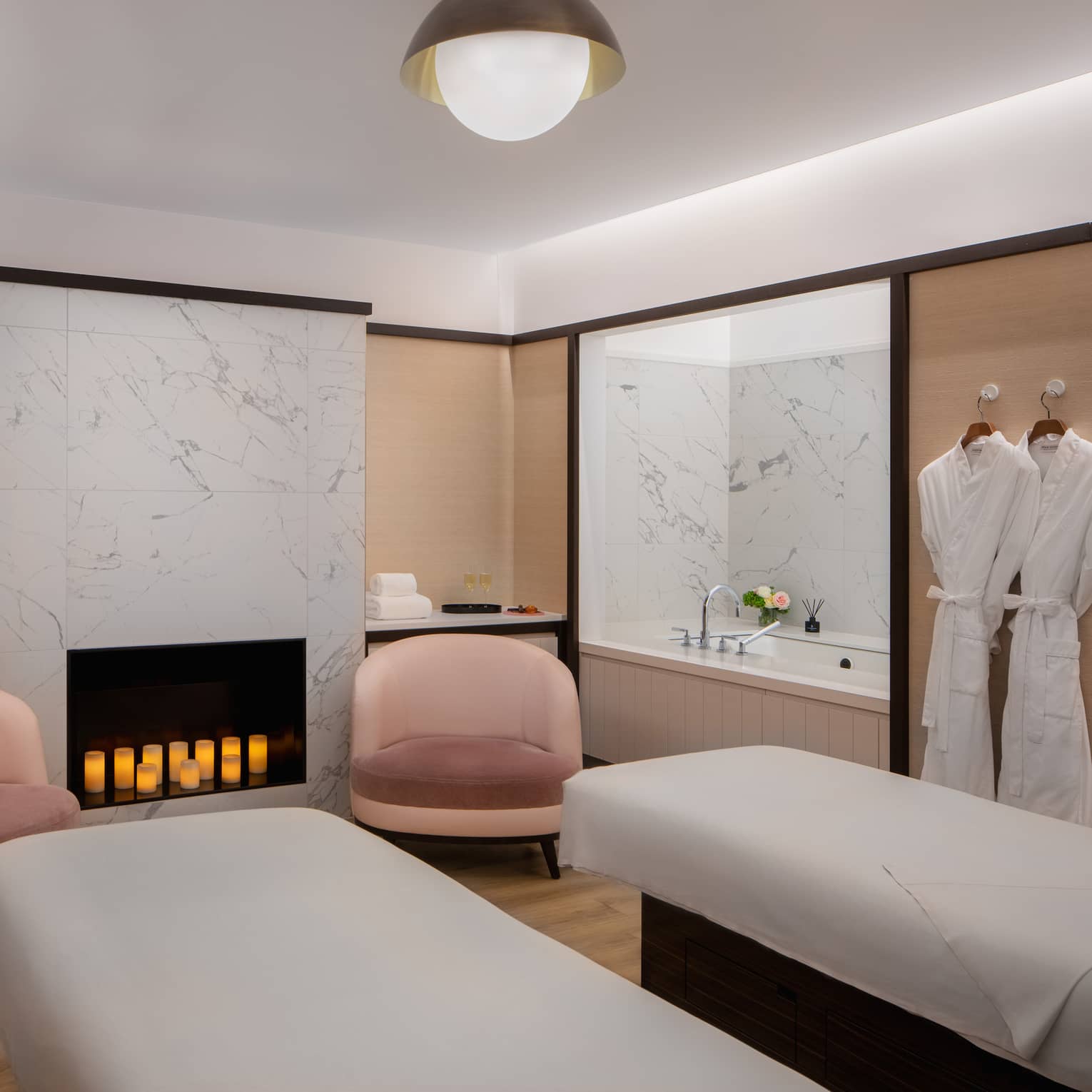 Couple's spa suite with a white-tiled fireplace filled with lit candles, an inset bathtub, two pink arm chairs, two side-by-side treatment tables and a pair of white robes