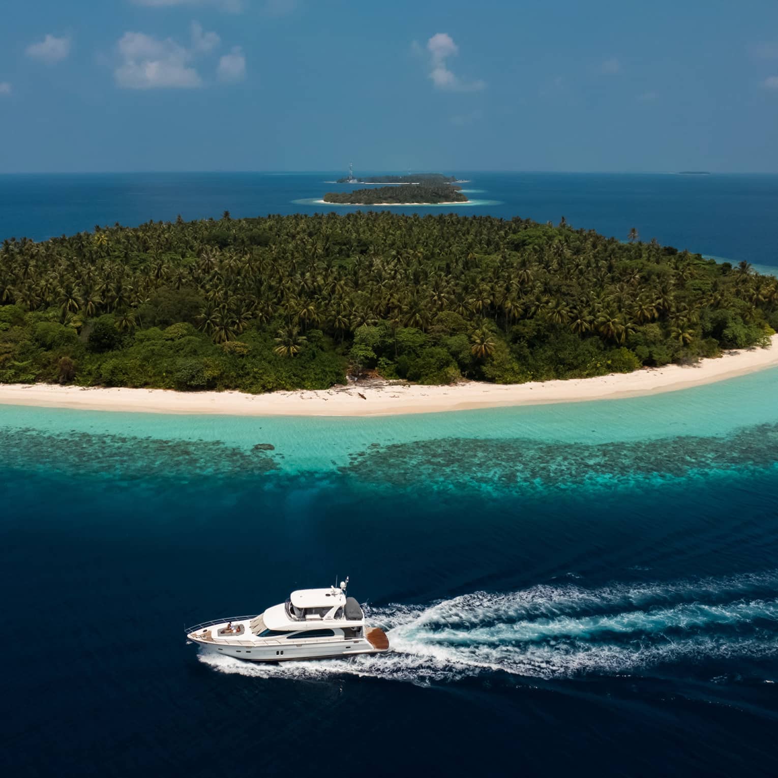 On a calm ocean, a yacht cruises past a small island with dense vegetation and white sand beach, two similar islands beyond.
