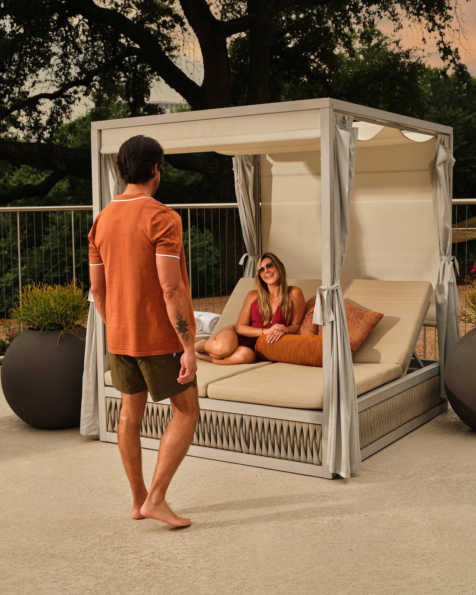A man walks towards a woman lounging in a cabana surrounded by large potted plants