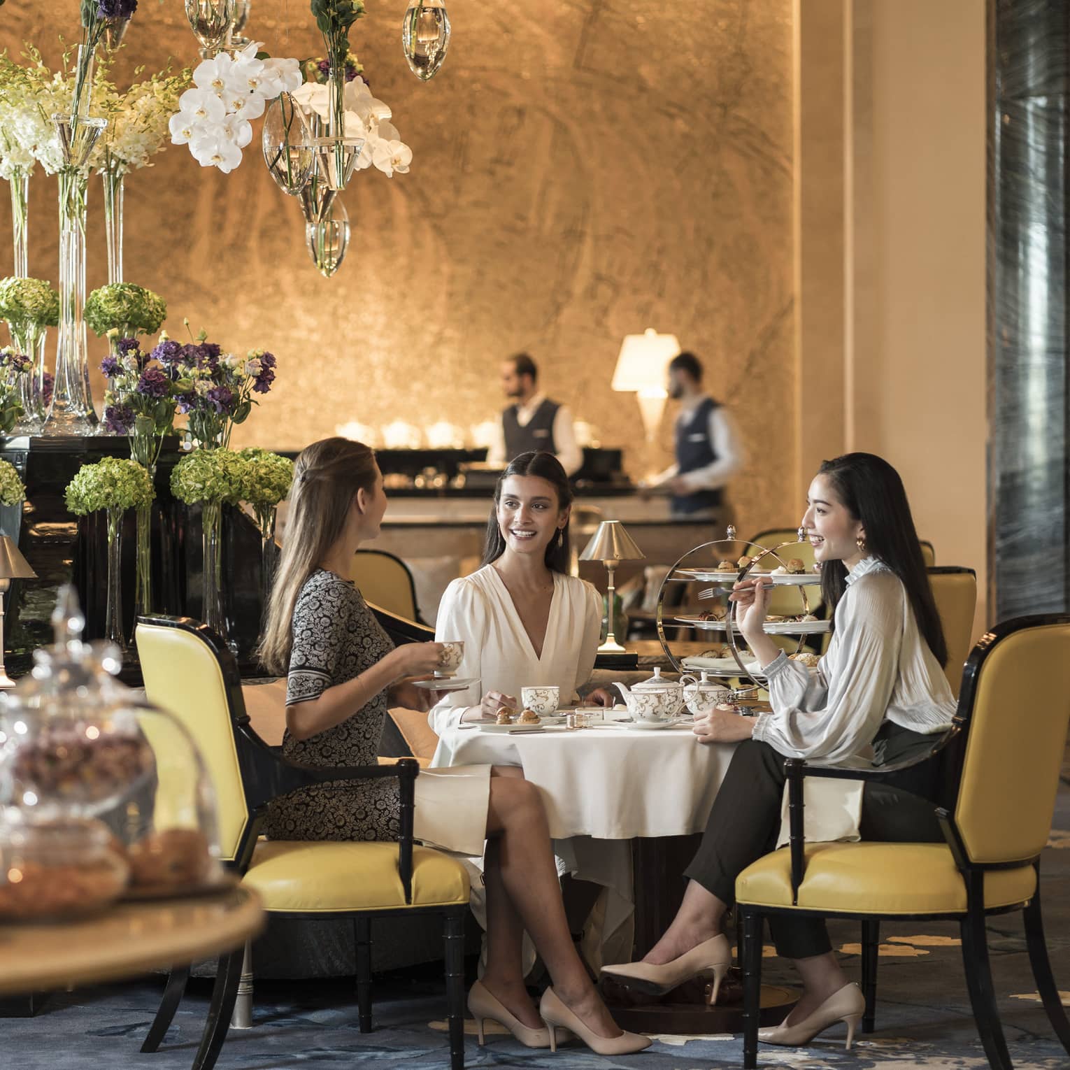 In an elegant restaurant, three smartly dressed diners enjoy an afternoon tea beside a feature of tiered flower-filled vases.