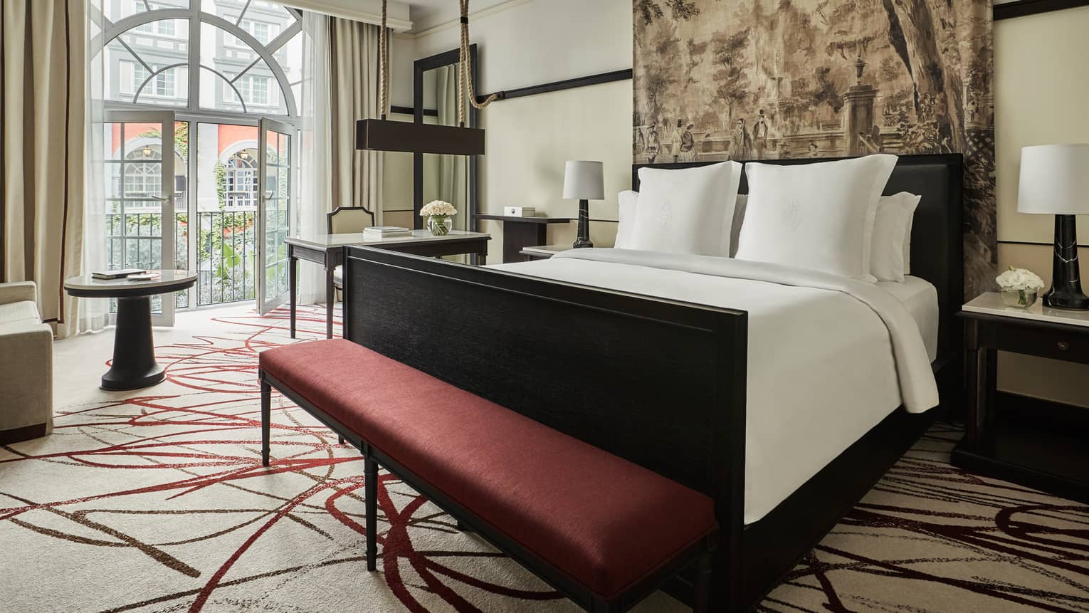 Premier Room bed under print headboard, red bench at foot, sunny arched balcony glass doors
