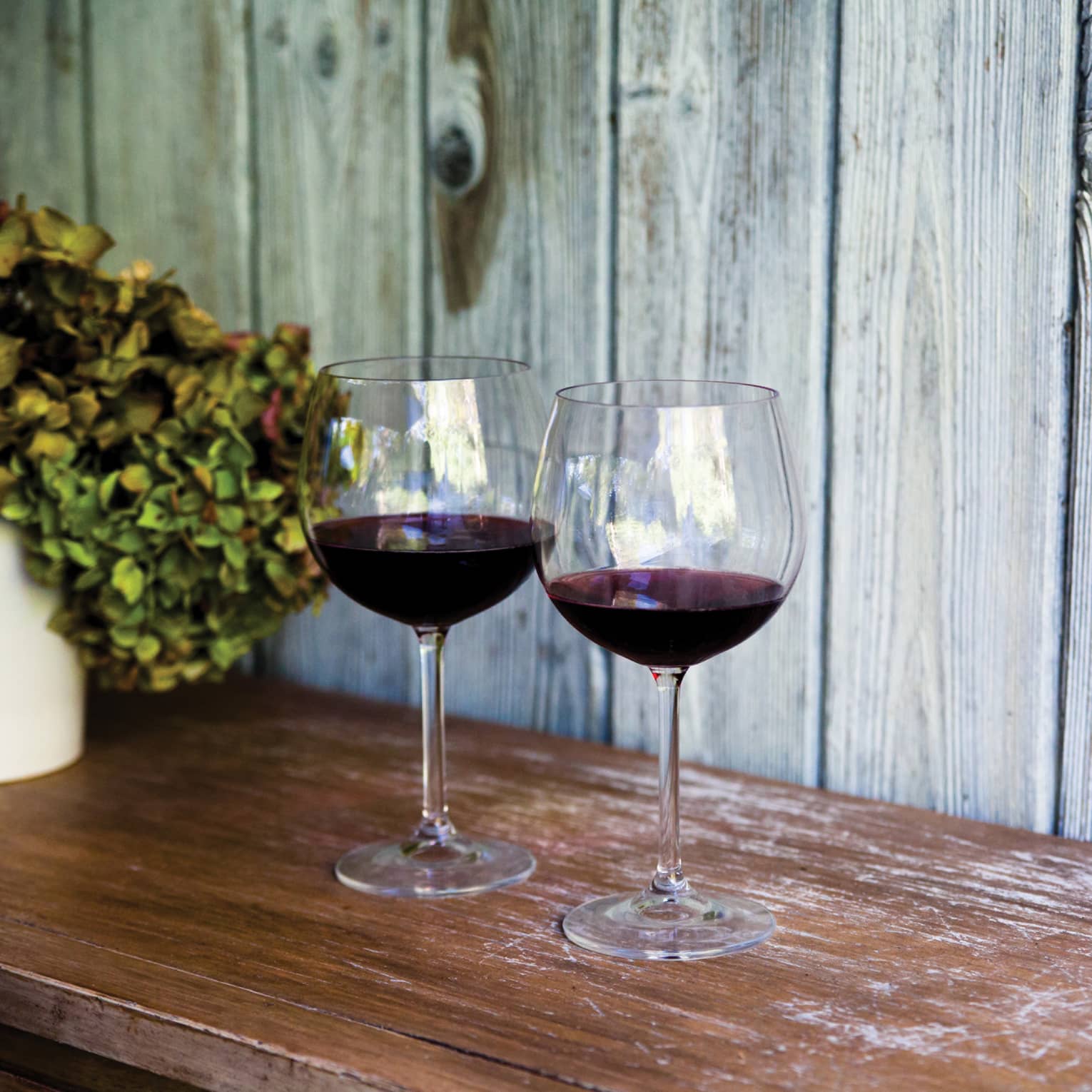 Two glasses of red wine and sit on wooden table, with white vase filled with hydrangeas 