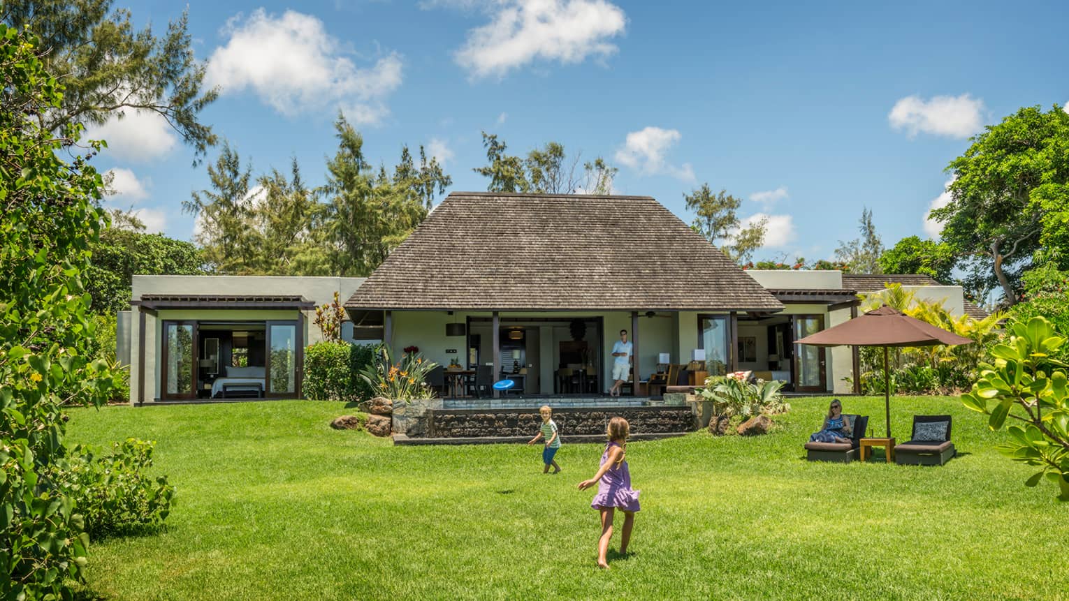 Young children play on green lawn in front of bungalow as dad watches from covered patio