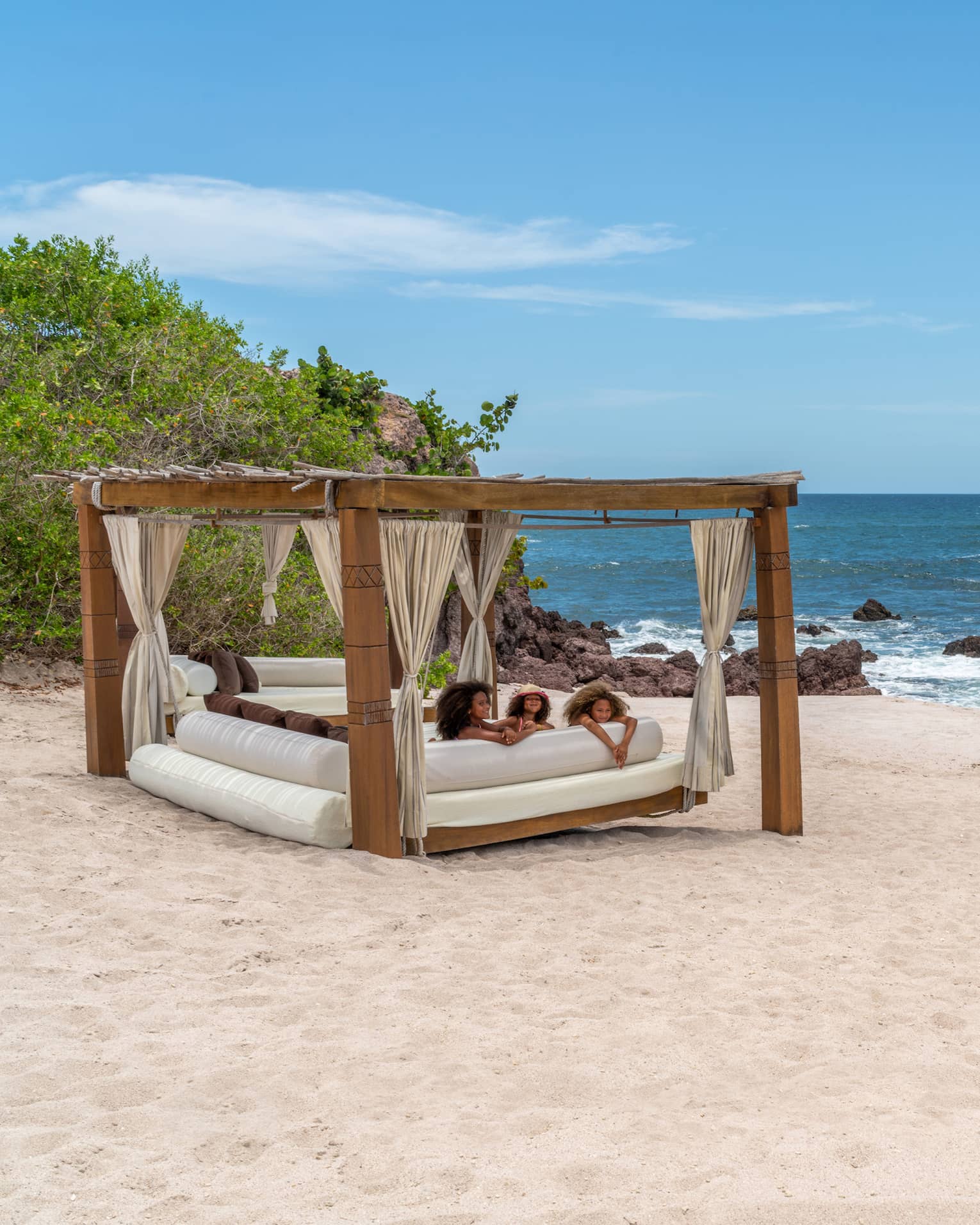 A cabana with three children laying on a plush seat underneath it on the beach, the ocean and plants are in the background.