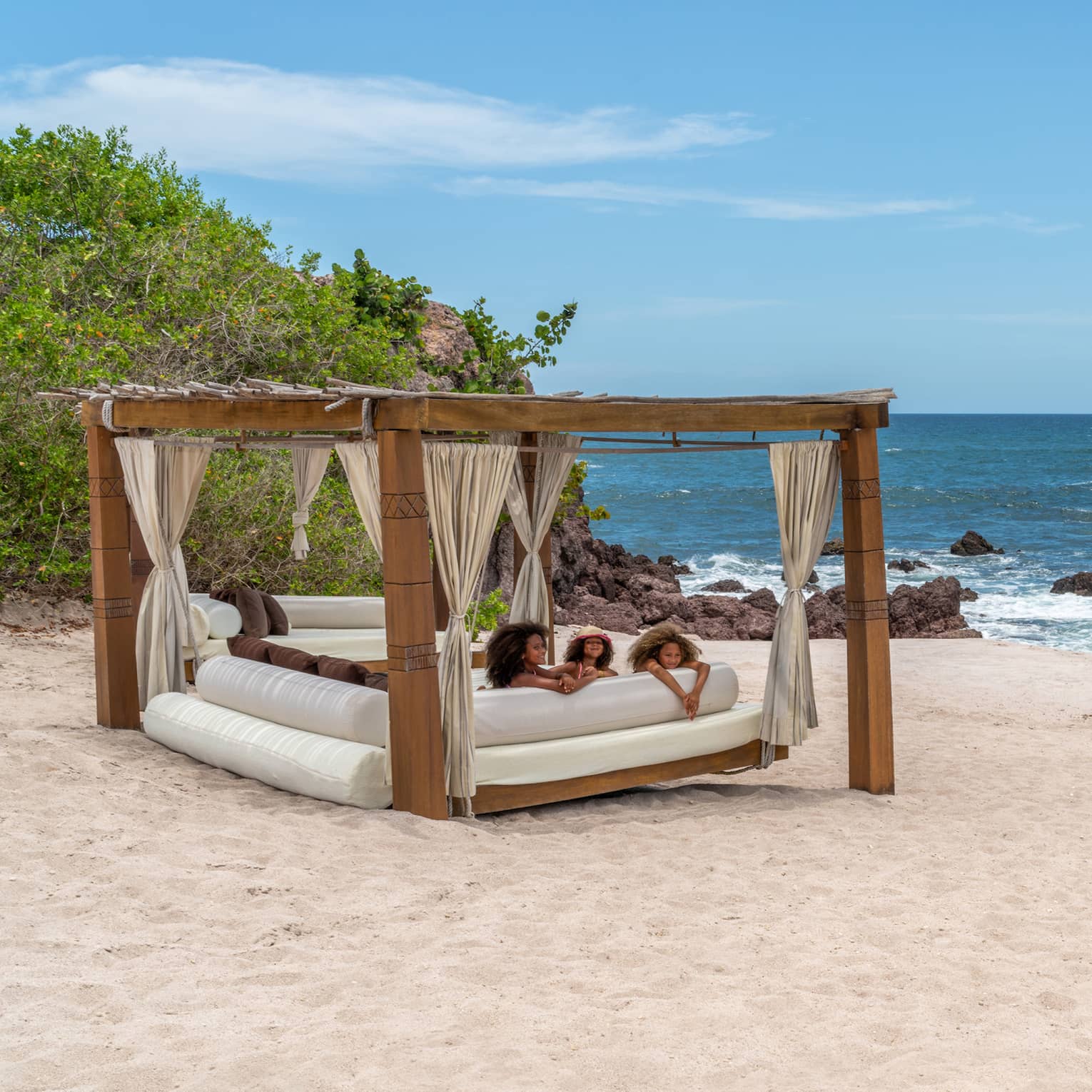 A cabana with three children laying on a plush seat underneath it on the beach, the ocean and plants are in the background.