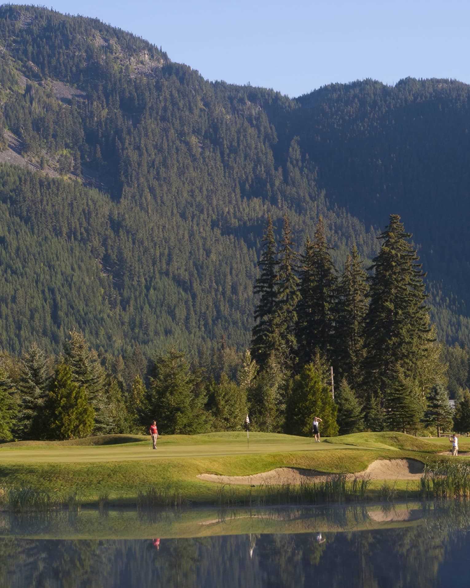 Distant golfers on a green, a pond in front, evergreens behind, dwarfed by a treed mountain filling most of the frame beyond.
