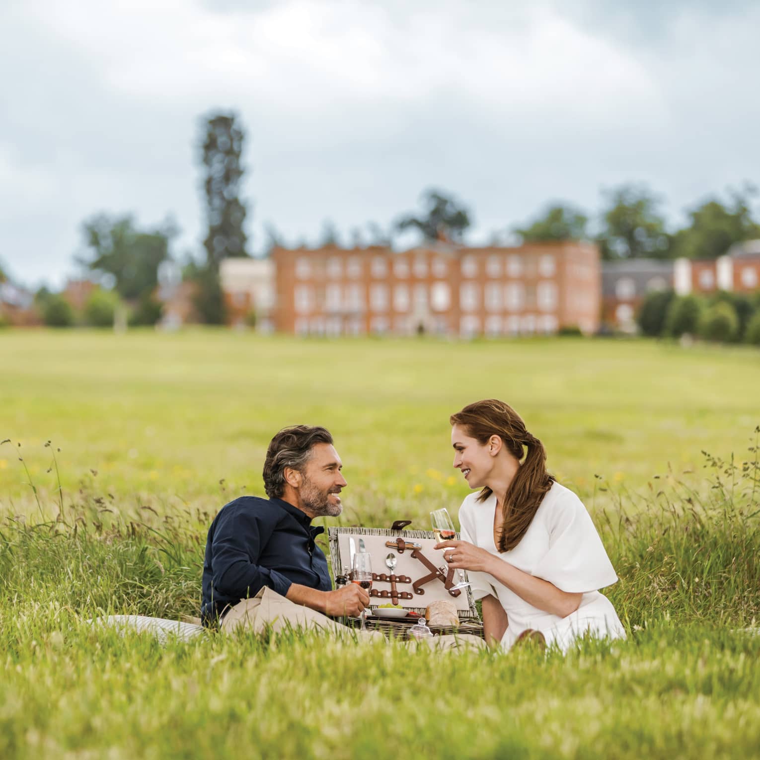 Couple with wine glasses lies in tall green grass with open picnic basket, hotel manor in background