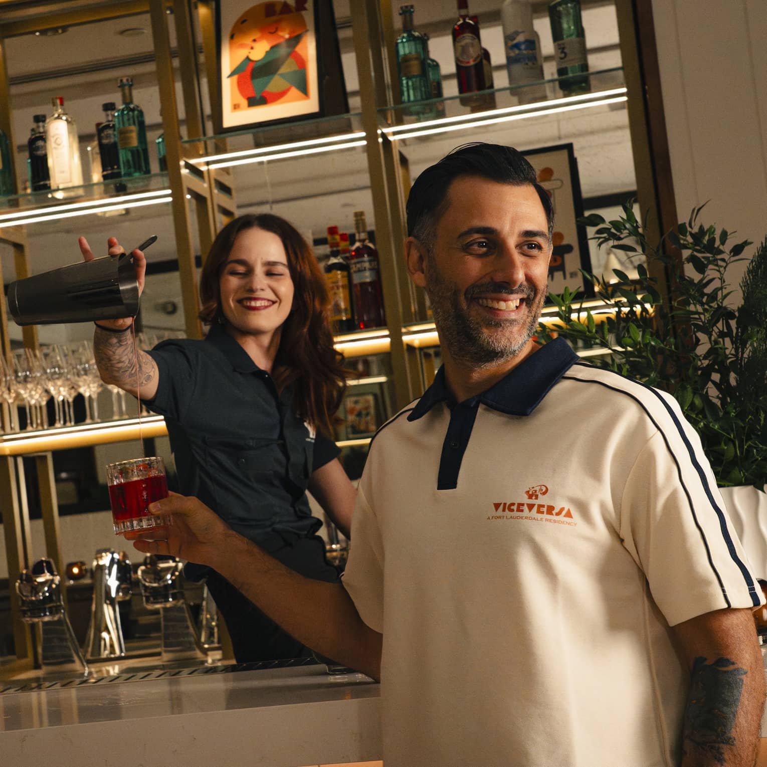 Smiling bartender pours a drink into a glass being held by another smiling person