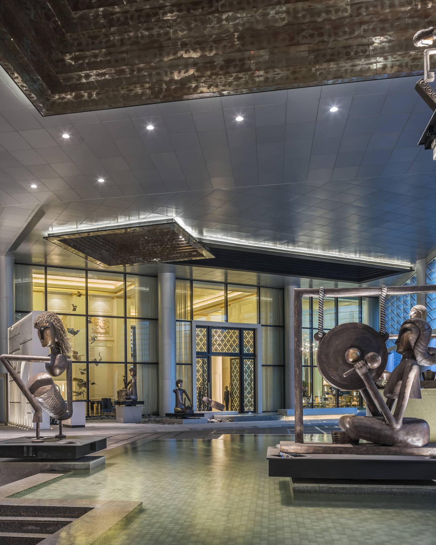 Hotel entrance with large modern Indonesian statues in fountain, doorman holding open glass doors in background