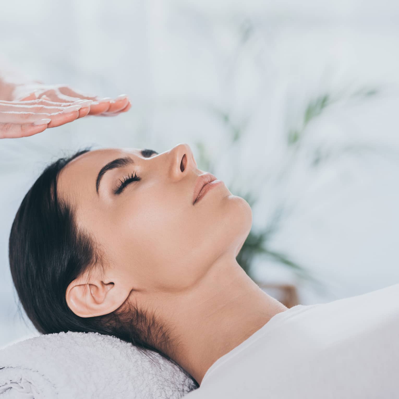 Woman with eyes closed lays on rolled up towel on spa table under therapists' hovering hands