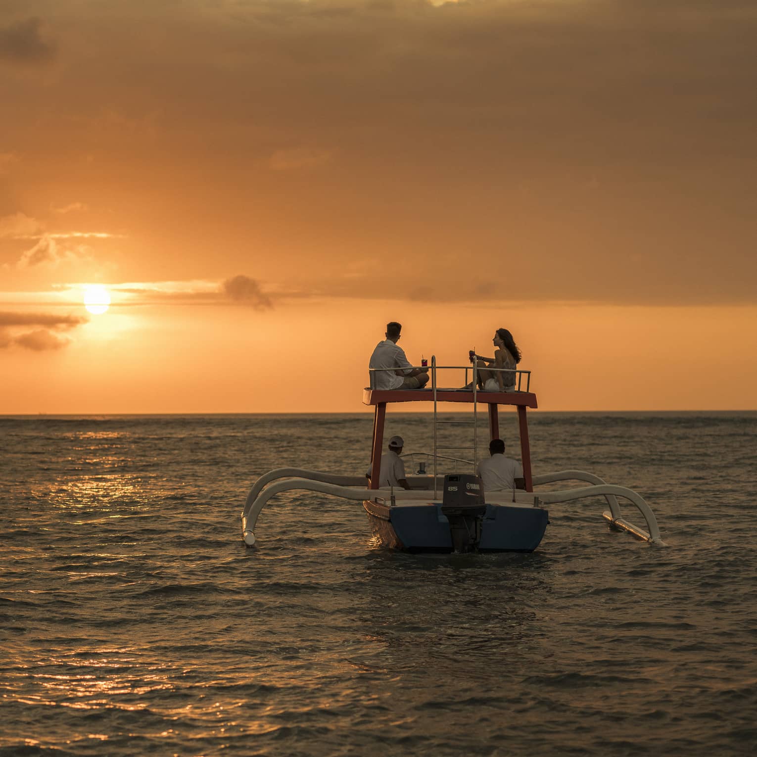 Couple sits on roof of boat sailing on ocean at sunset