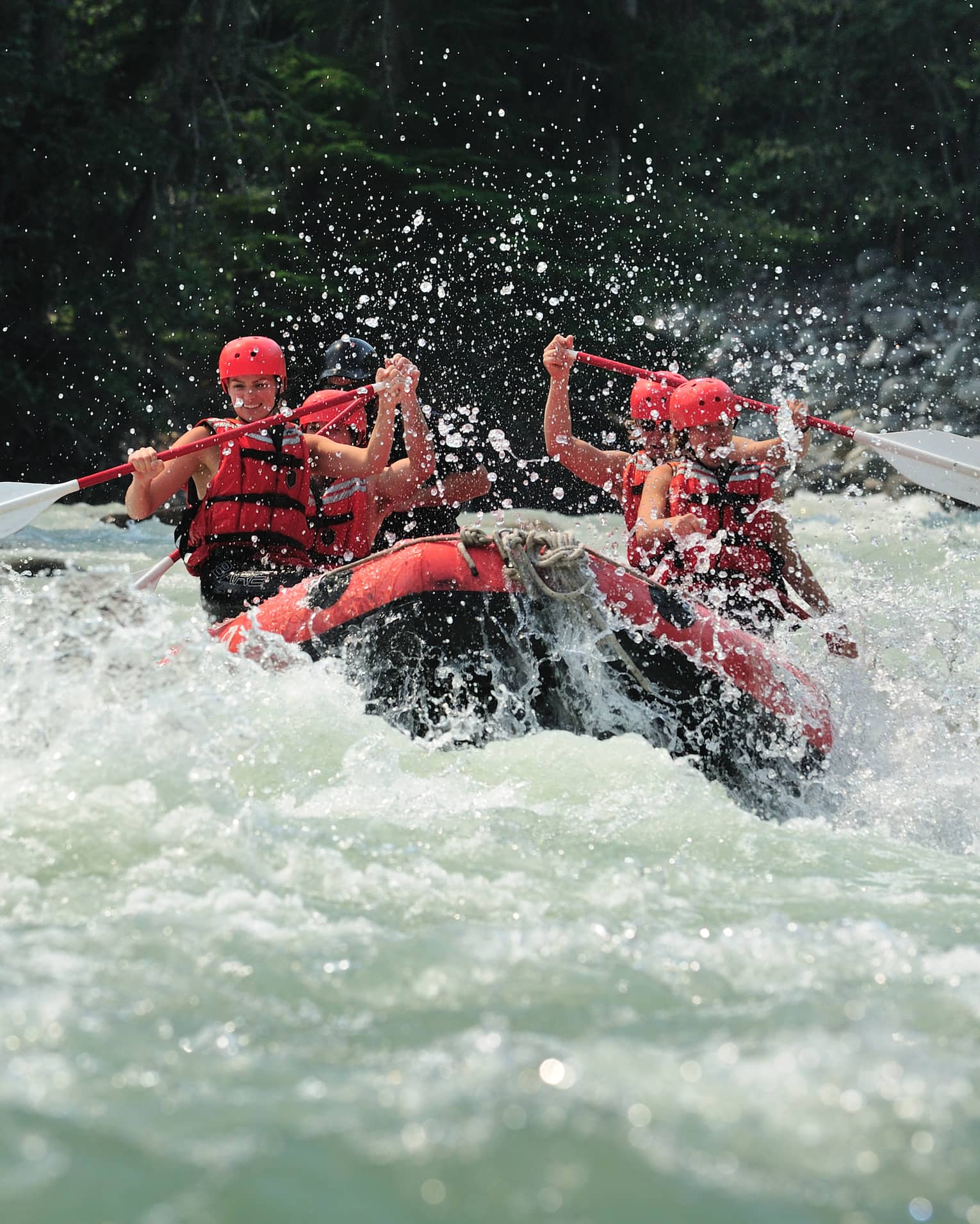 Smiling paddlers in red helmets and lifejackets steer an inflated raft through raging white water, passing evergreen trees.