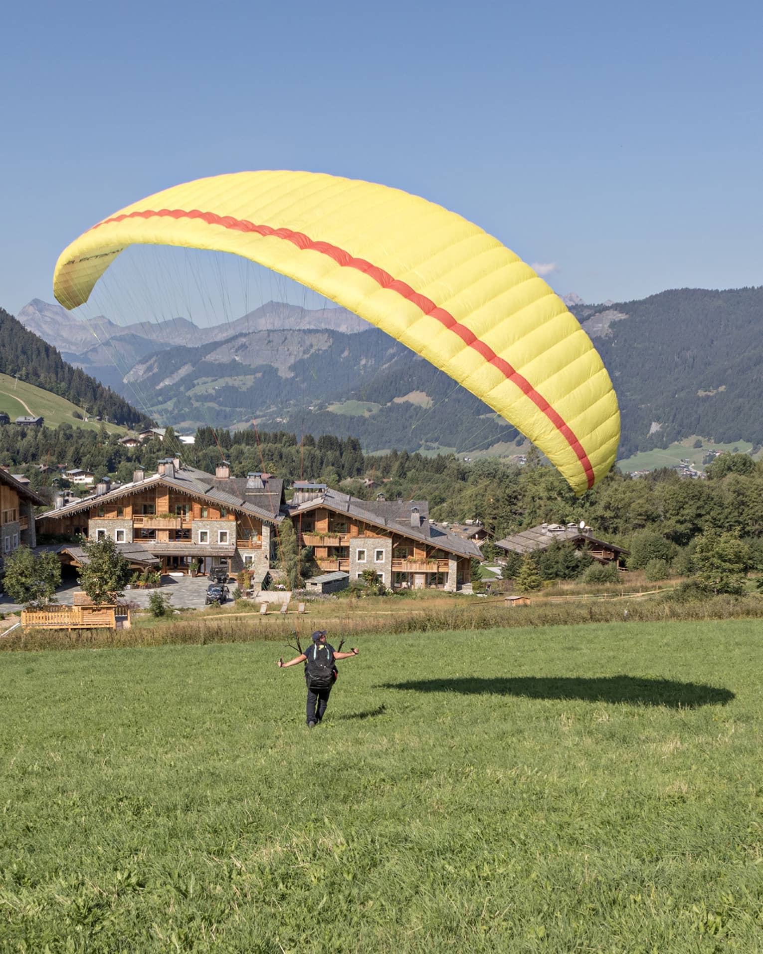 Person on meadow holding yellow parachute in air, facing rustic Megève hotel and verdant mountains
