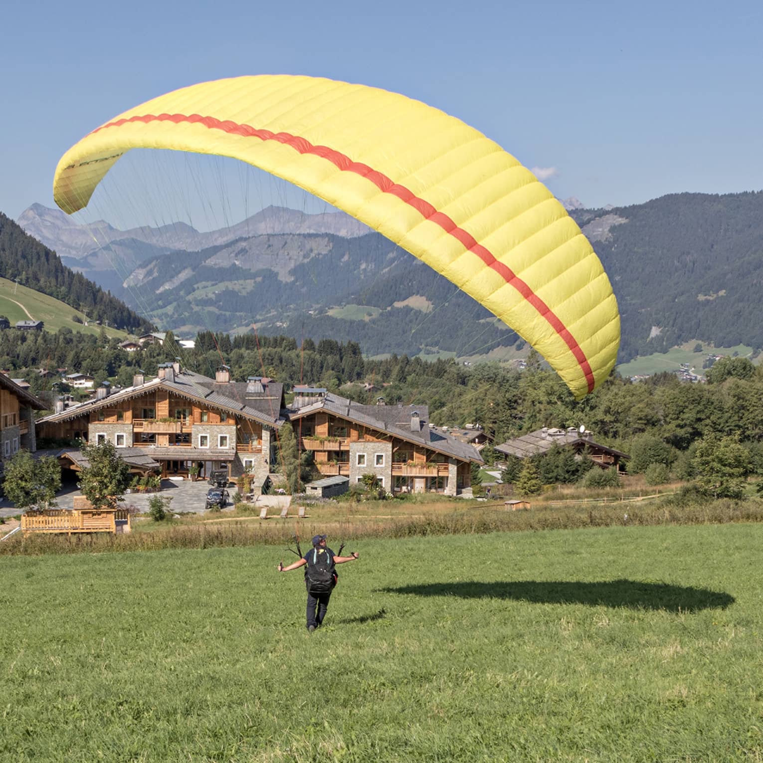 Person on meadow holding yellow parachute in air, facing rustic Megève hotel and verdant mountains