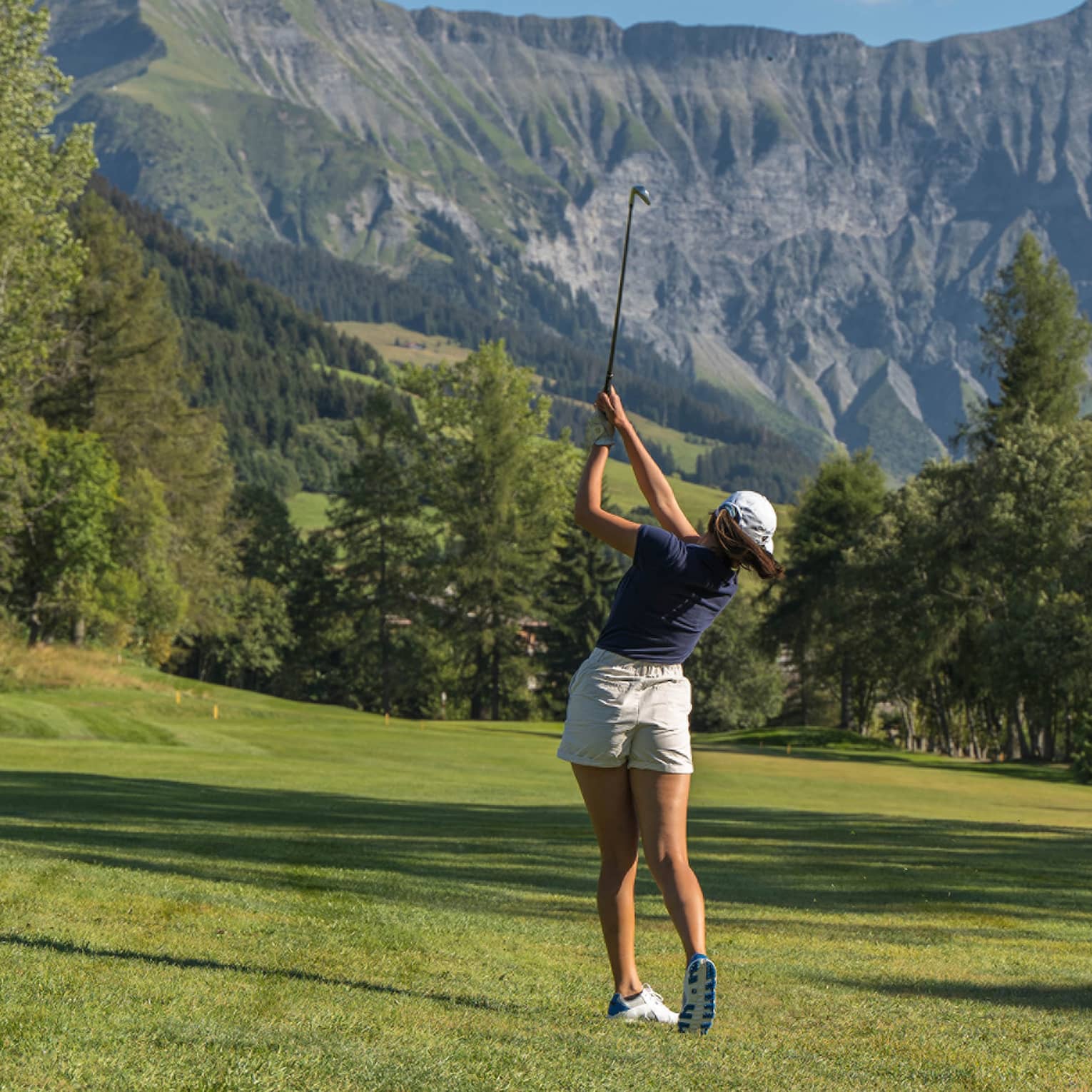 A golfer taked a swing on a golf course on a beautiful sunny day. A mountain covered in greenery is in the background.