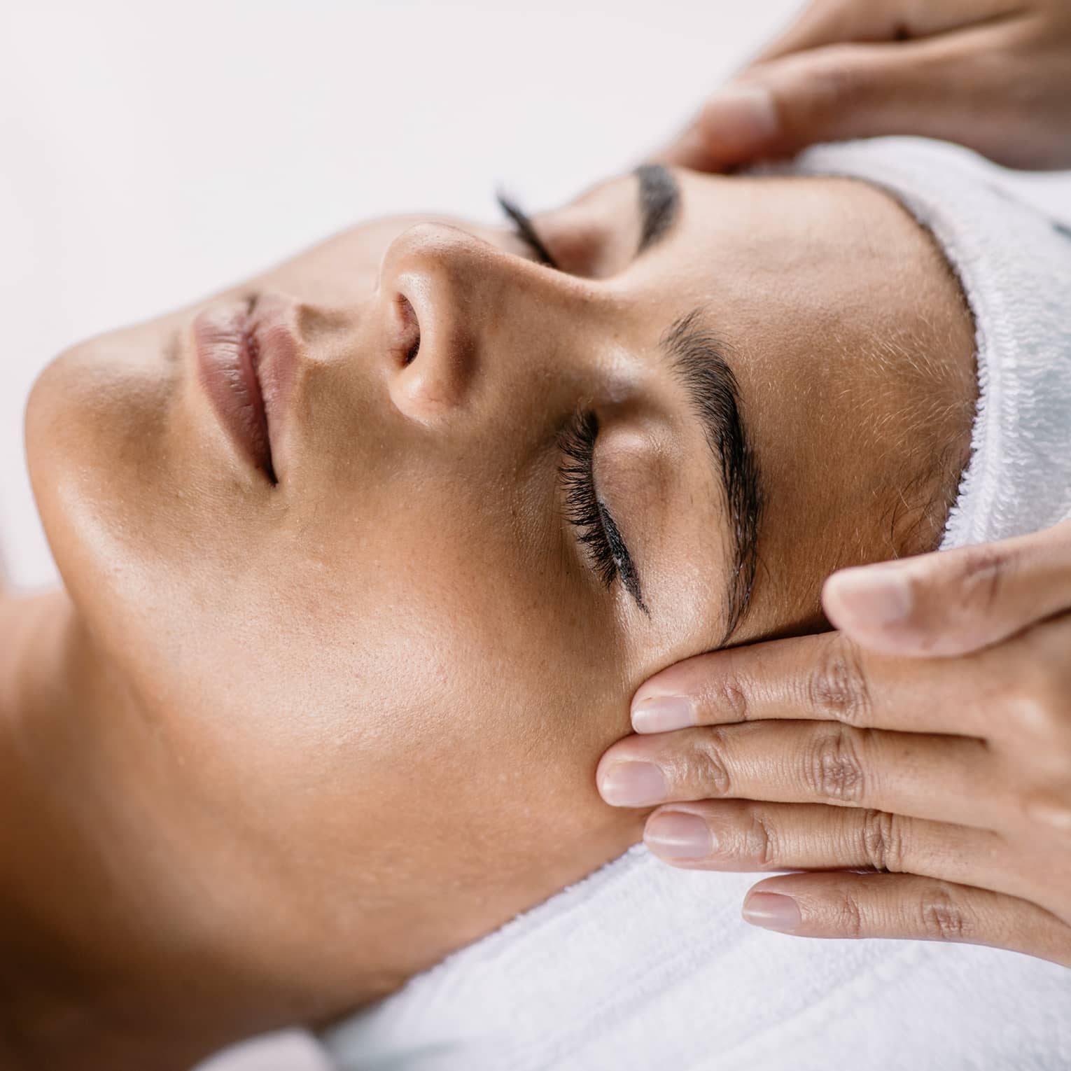 Hands massage woman's temples as she closes her eyes, spa towel around hair