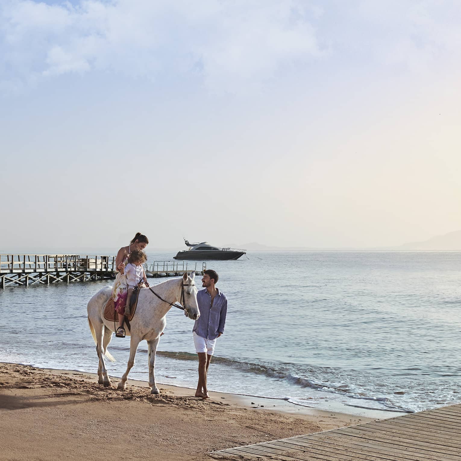 An adult and child on a white horse on a beach, led by another guest, with a wooden pier and a yacht visible in the distance.