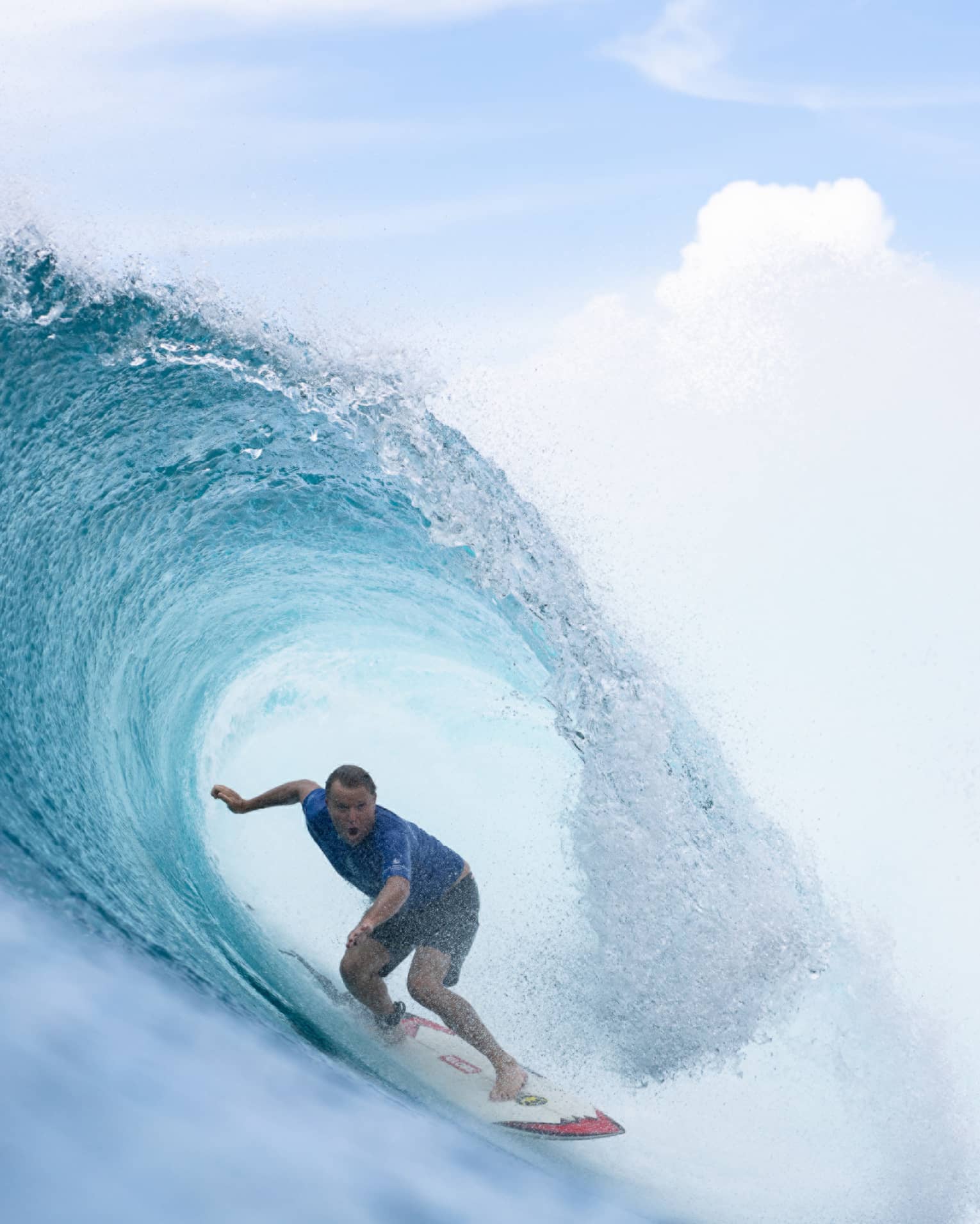 Close-up of a rippling, crystal-clear wave curling over an open-mouthed surfer crouched on a board under a cloudy sky.