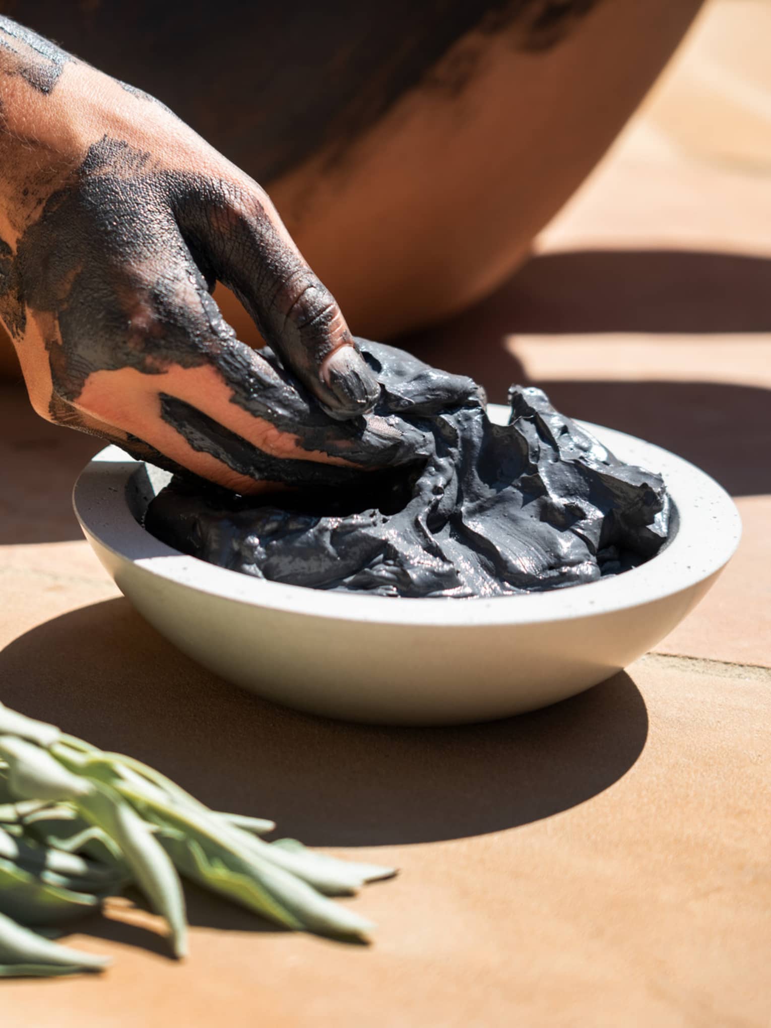 Hand covered in black clay reaching into a bowl of spa mud next to sage bundle